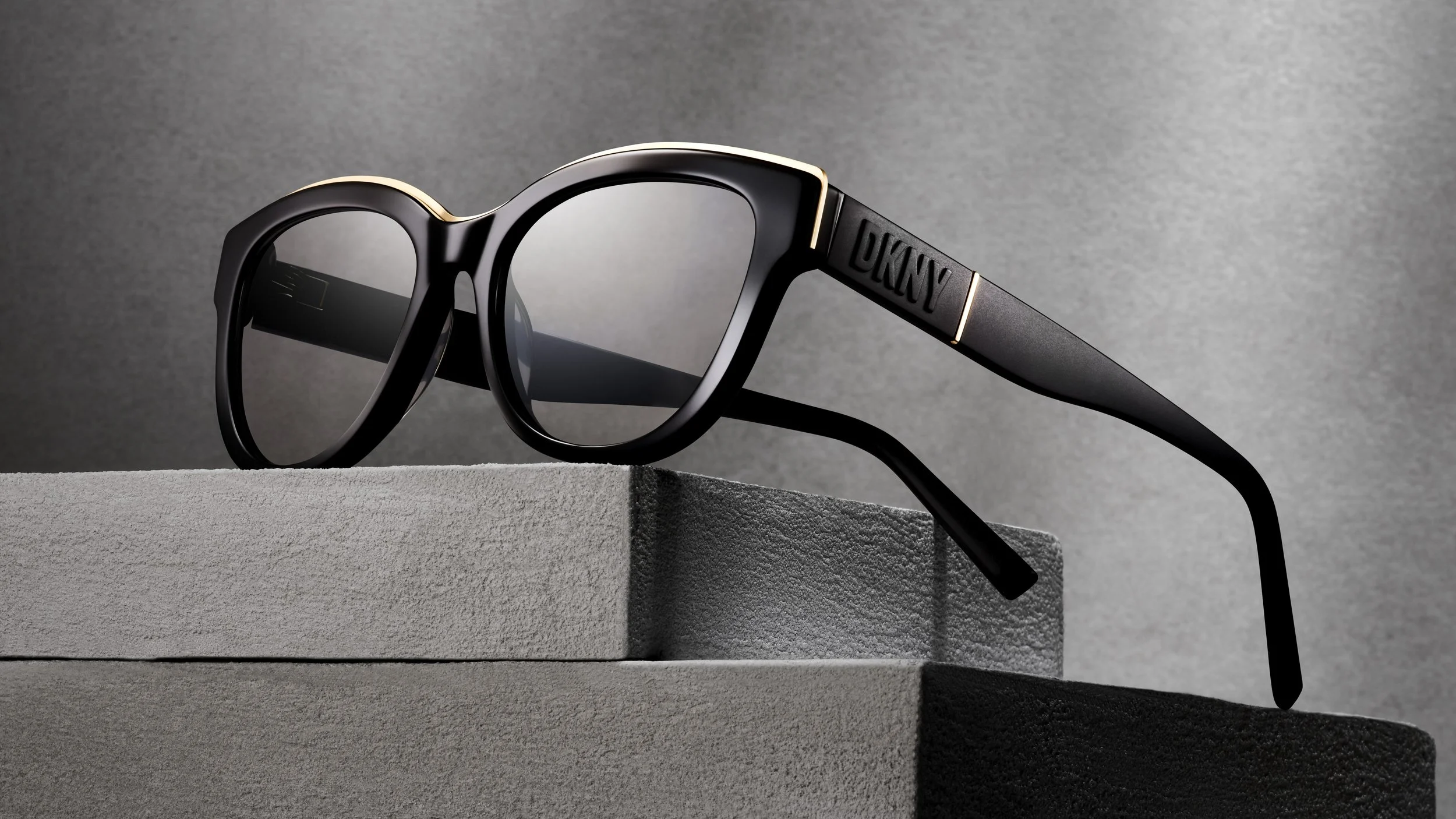 Close-up of black sunglasses with gold accents resting on a gray textured surface, with a gray background.