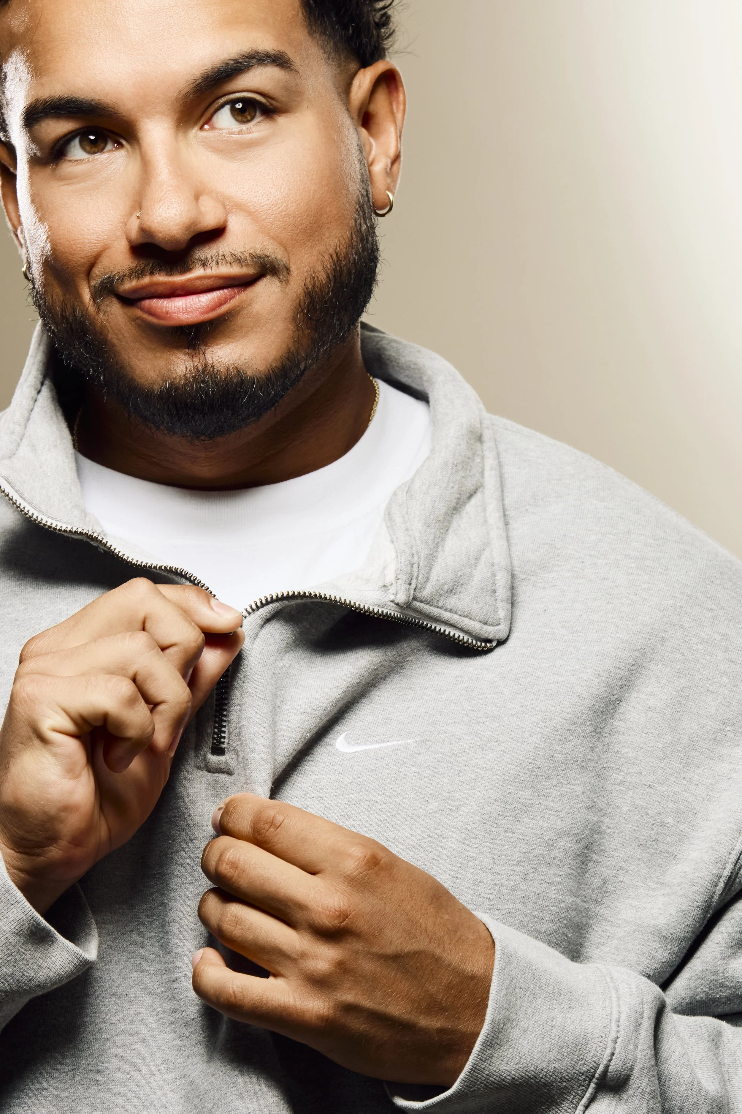 A man with a beard, earrings, and a nose piercing, wearing a light gray Nike sweatshirt, zipping up the sweatshirt.