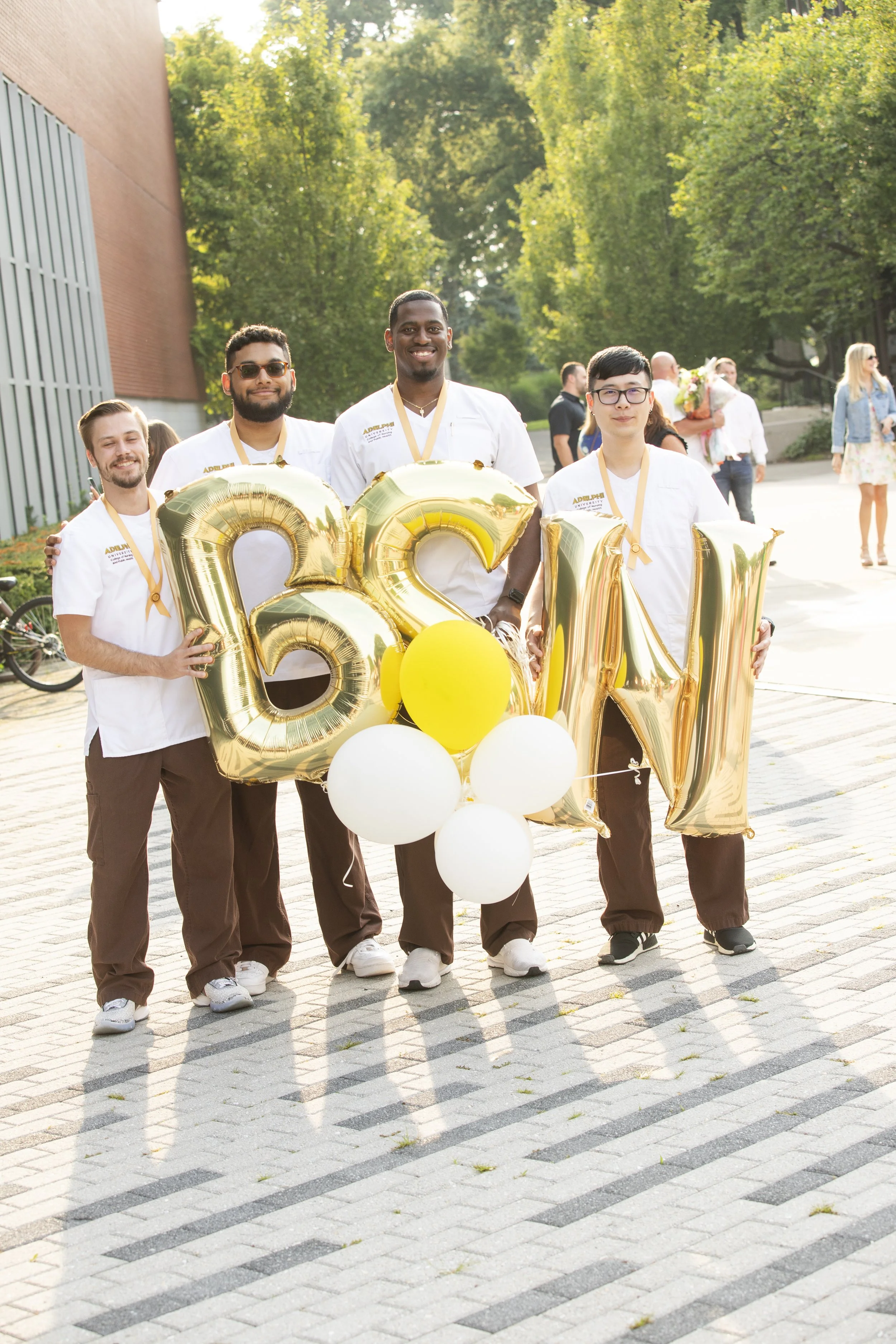 A group of five smiling people holding large gold balloons spelling 'GWN' and yellow and white balloons, standing outdoors on a sunny day with trees and people in the background.