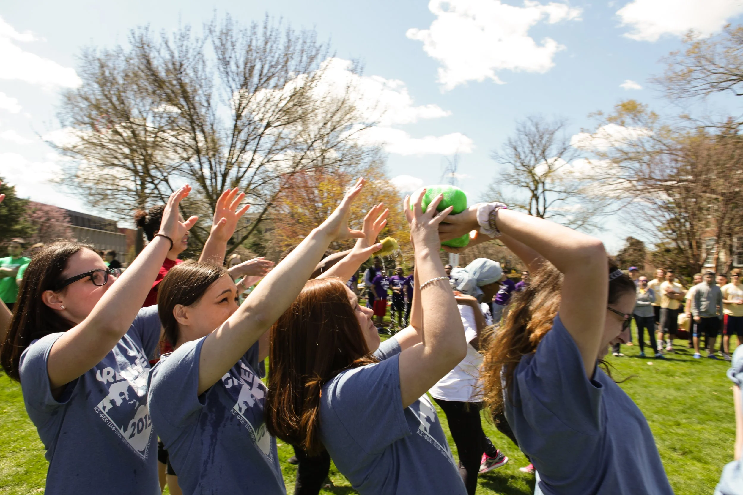People participating in an outdoor event, throwing a green foam object into the air on a sunny day with trees and a crowd in the background.