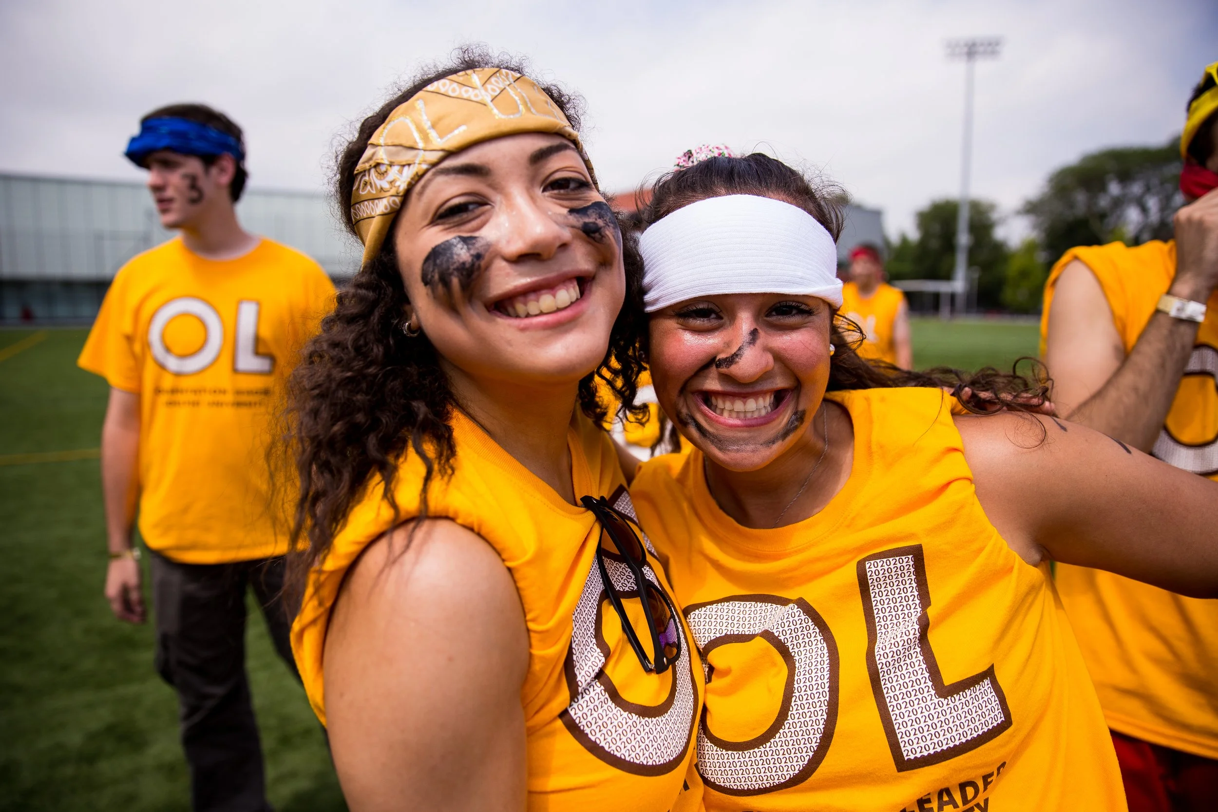 Two smiling women wearing yellow shirts and white headbands, celebrating on a sports field with other people in the background.