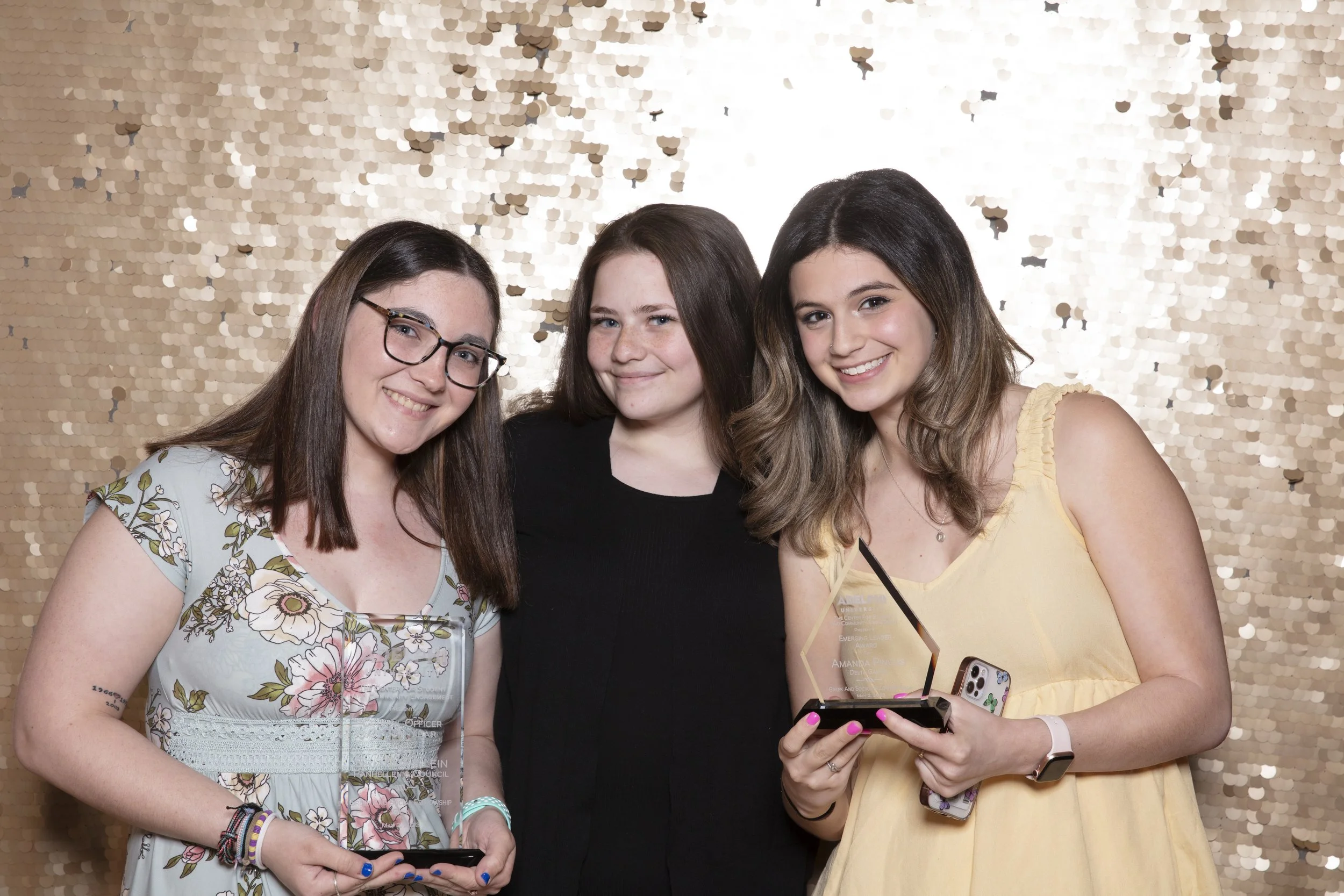 Three young women smiling, one holding a trophy, standing in front of a gold sequin backdrop.