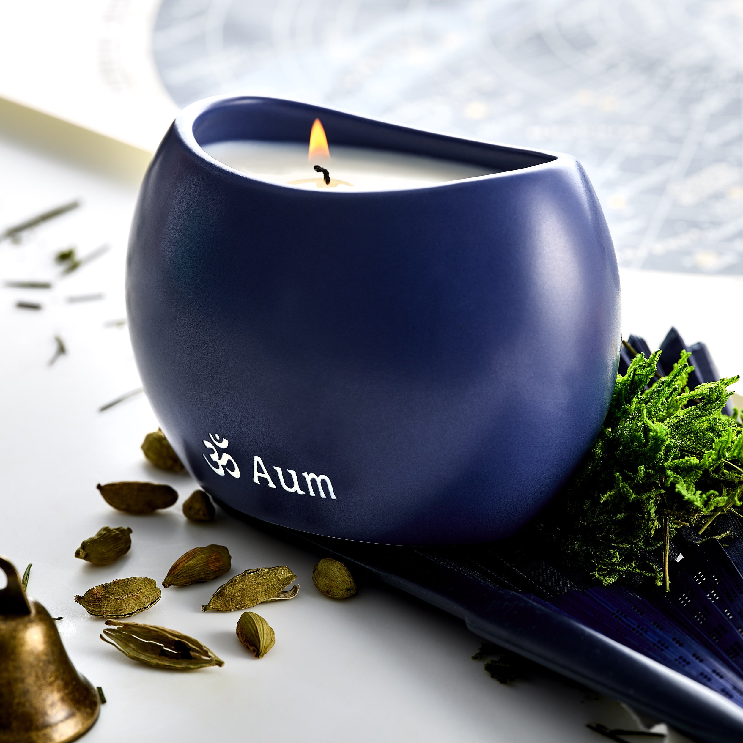 Close-up of a blue candle with a small flame, surrounded by cardamom and coriander seeds on a white surface, with green moss and a metallic object in the background.
