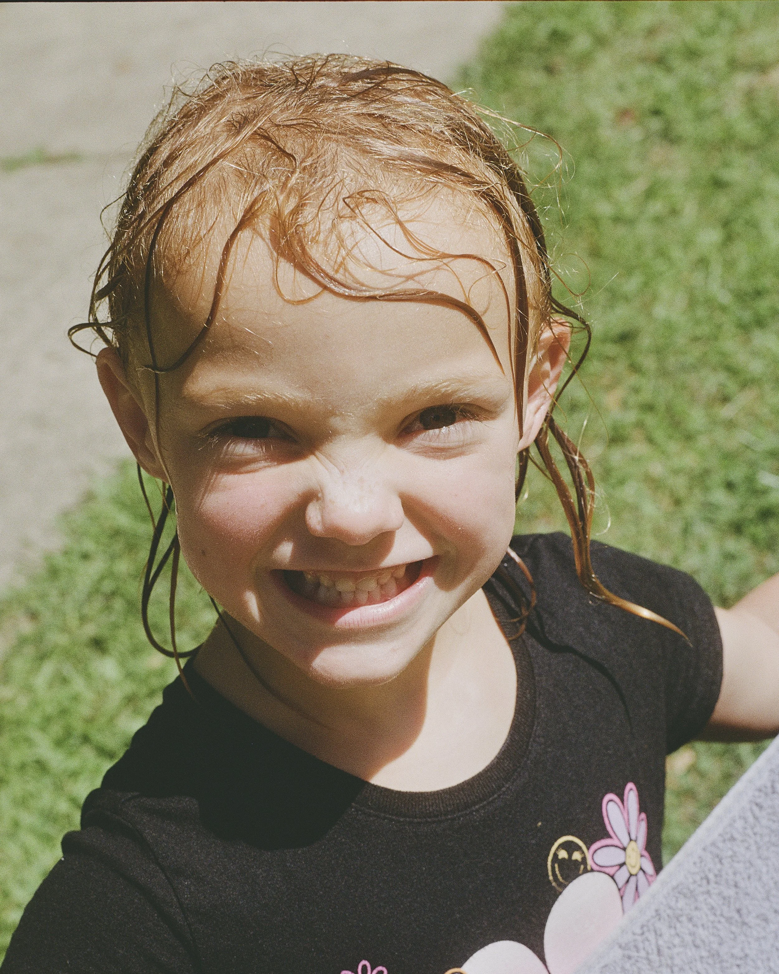 Close-up of a smiling child with wet, curly, red hair outdoors on a sunny day, wearing a black shirt with pink and yellow flower and smiley face designs, and standing on grass.