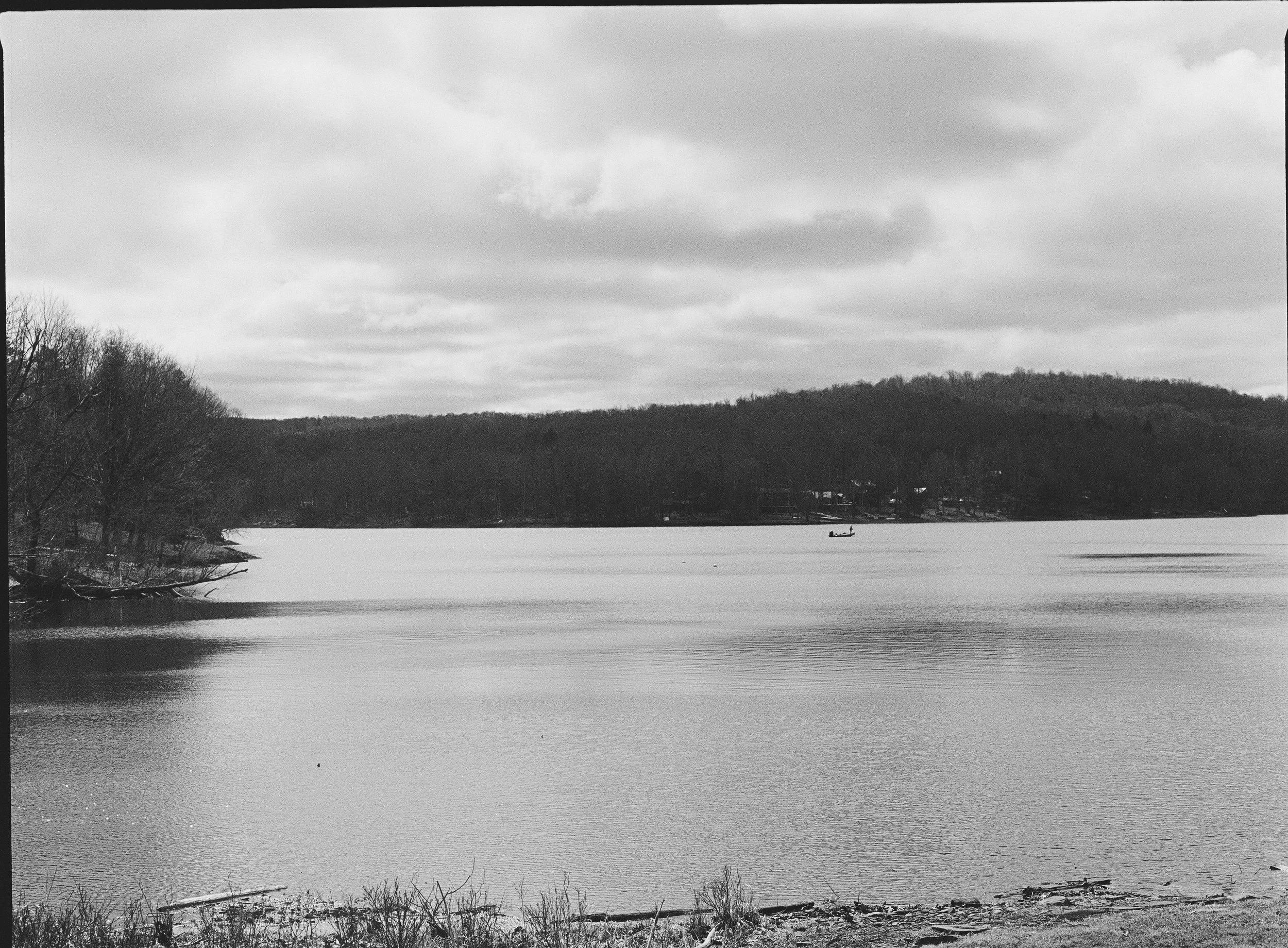 A black and white photo of a lake with a boat floating on the water and a hilly area with trees in the background.