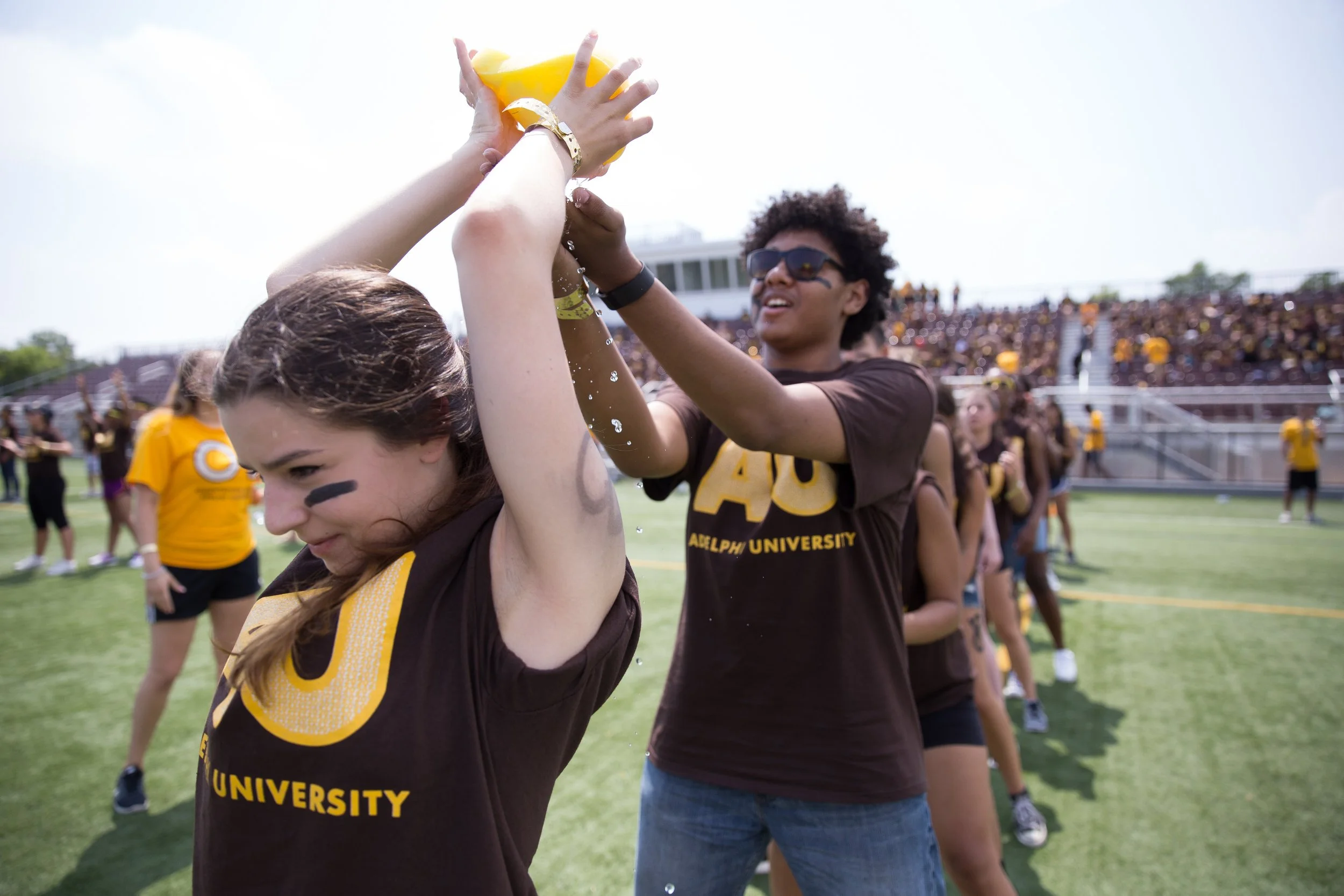 Students at Adelphi University participating in a water game on a sports field during a campus event, with a large crowd and stadium seating in the background.