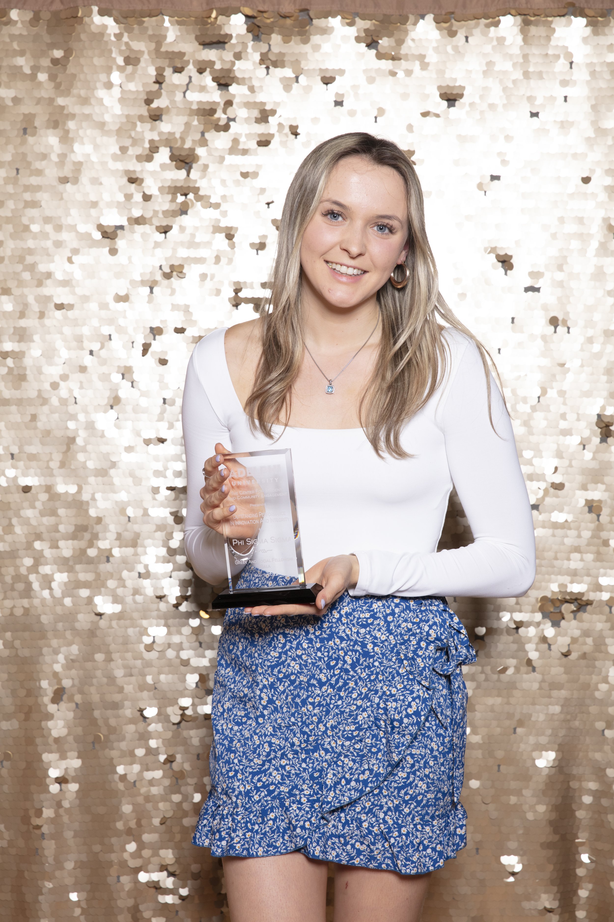 A young woman with blonde hair and blue eyes holds a glass award and smiles in front of a gold sequin backdrop.