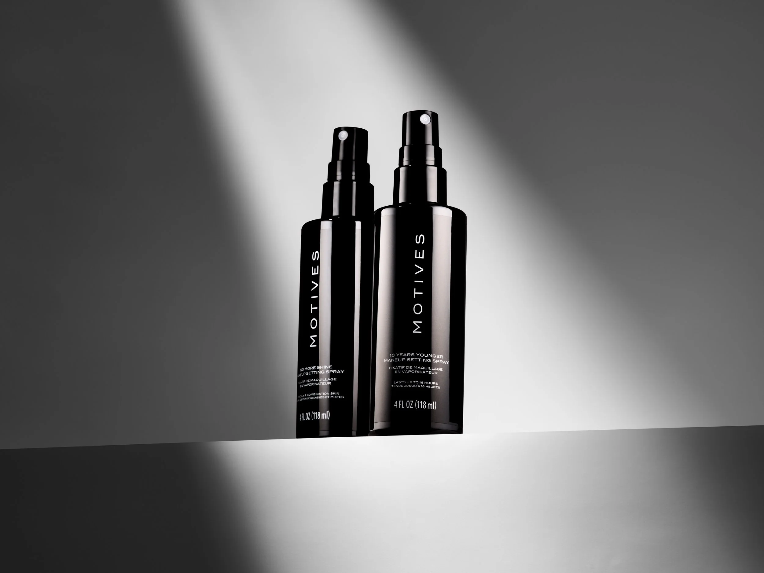 Two black bottles of MOTIVES makeup setting spray with spray nozzles, placed on a flat surface with dramatic lighting and a plain background.