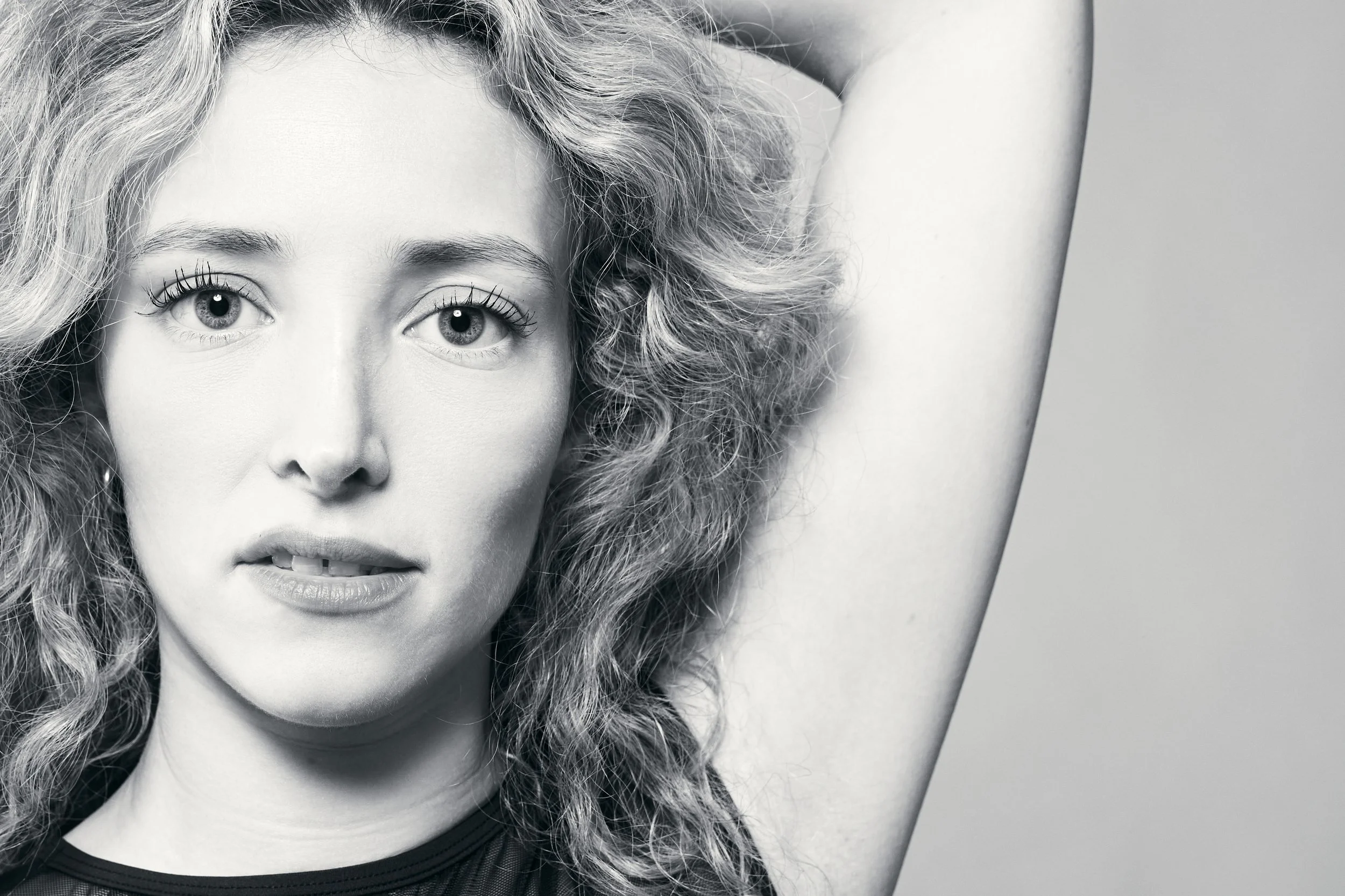 A close-up black and white portrait of a woman with curly hair, wide eyes, and her mouth slightly open, with one arm raised over her head.