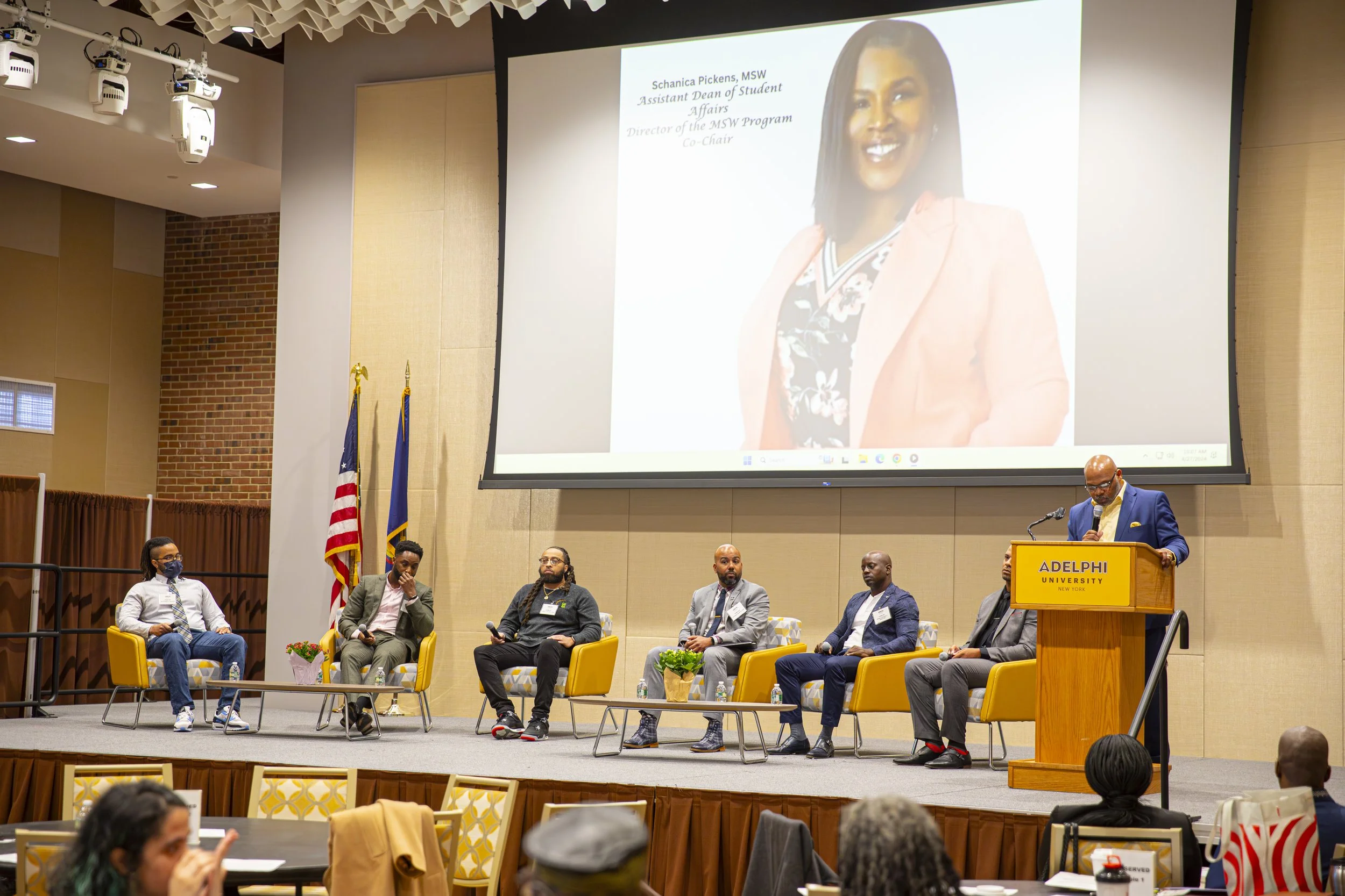 A panel discussion at Adelphi University, featuring six men seated on stage with a large screen behind them displaying a photo of a woman and her credentials. A man at a podium labeled 'Adelphi University' is speaking into a microphone. Audience memb