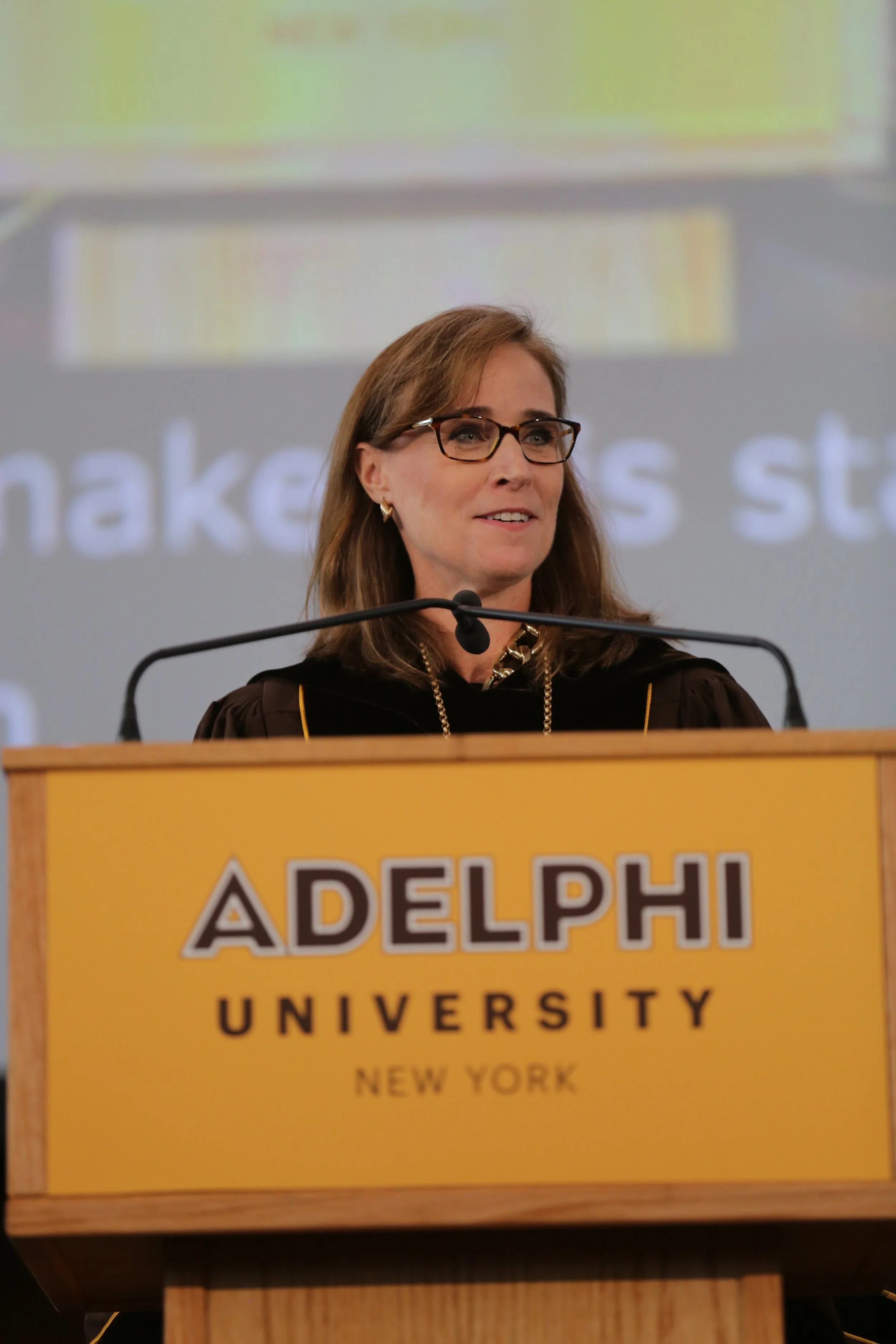 A woman with glasses and shoulder-length hair speaking at a podium with a microphone. The podium has a sign that reads "ADELPI UNIVERSITY NEW YORK." She is wearing a black academic gown and jewelry. In the background, a screen displays faint text and