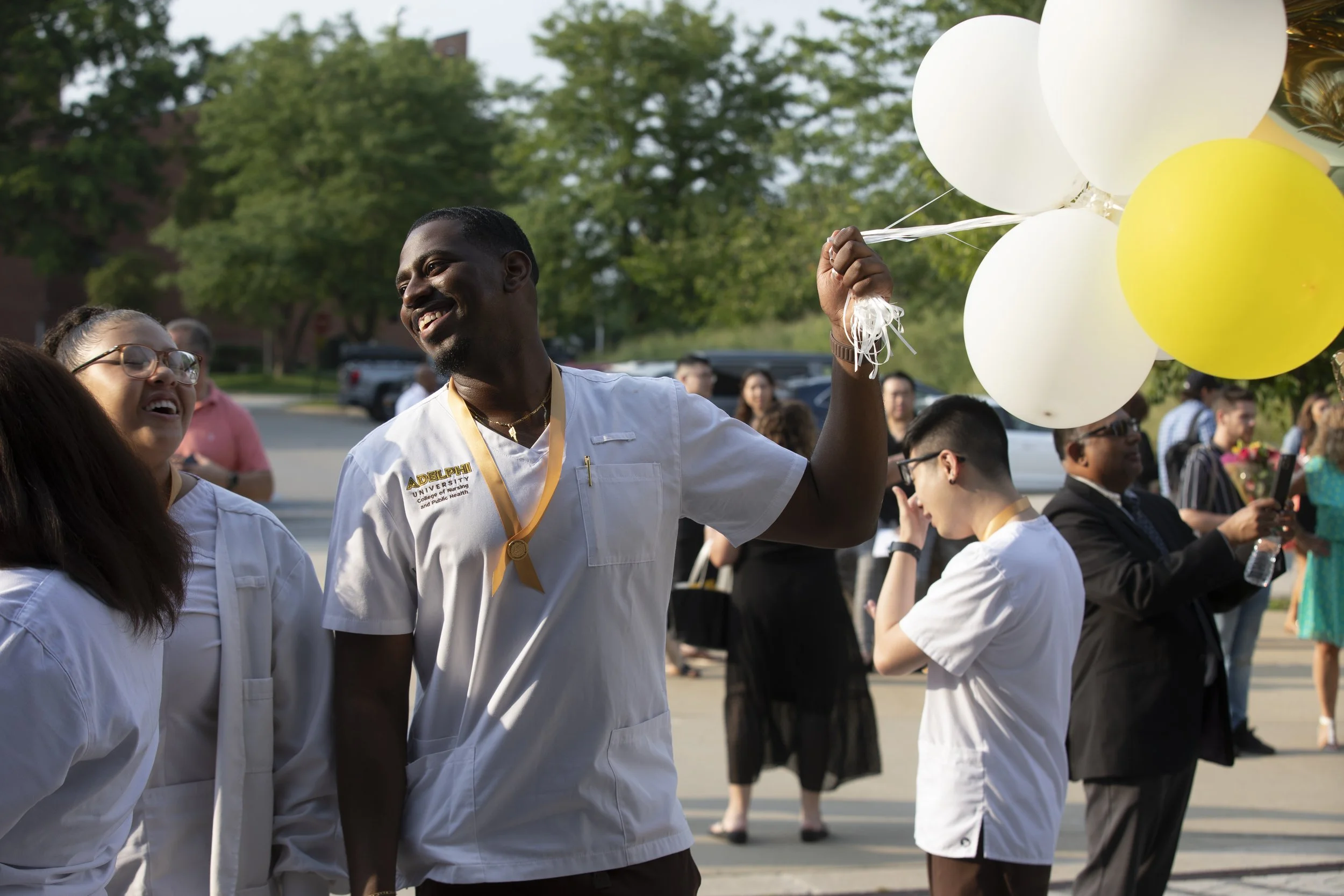 A graduation celebration with students in white gowns, smiling and holding balloons outdoors.