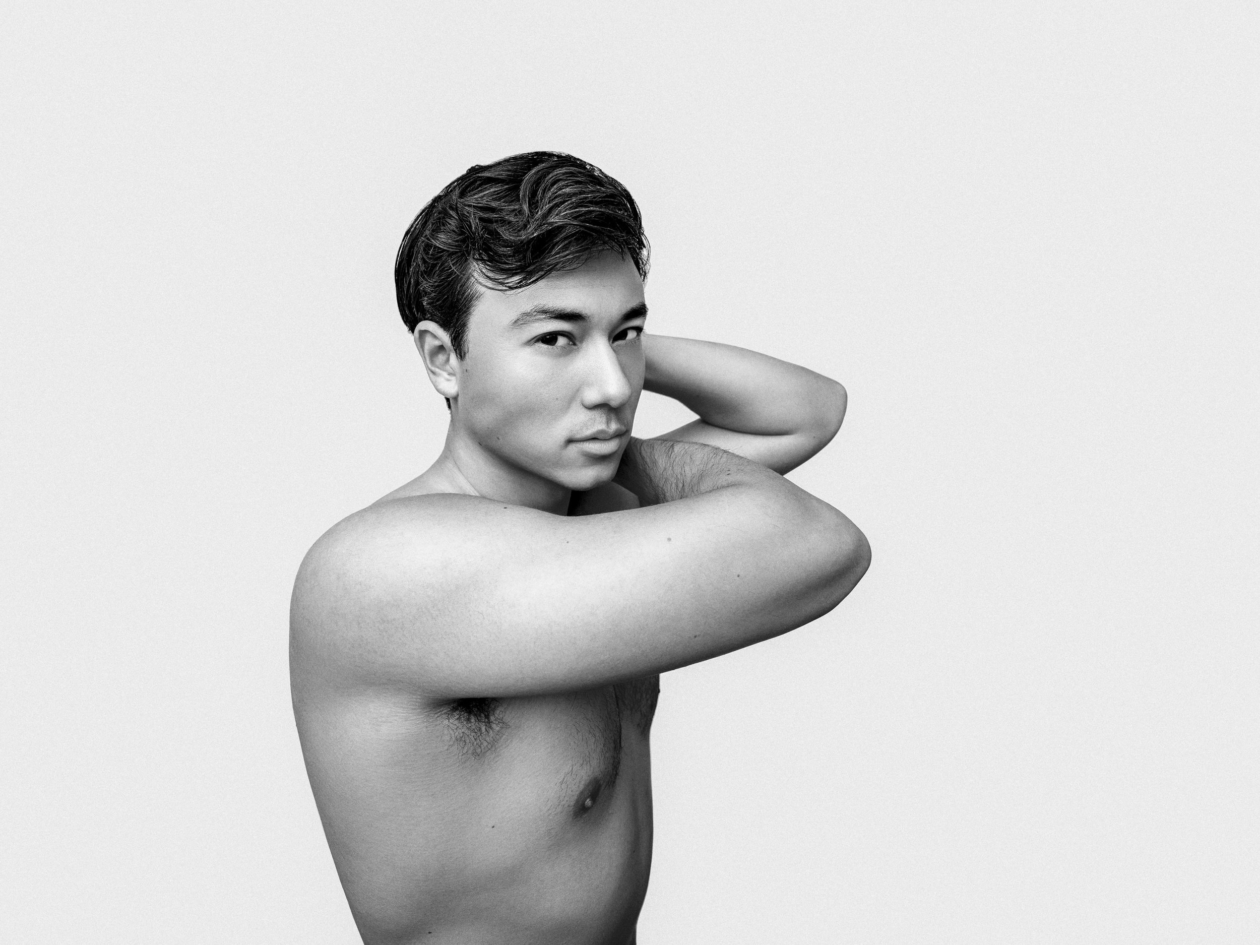 Black and white photo of a shirtless young man with dark hair, looking at the camera, with one arm bent and hand resting behind his head, standing against a plain light background.