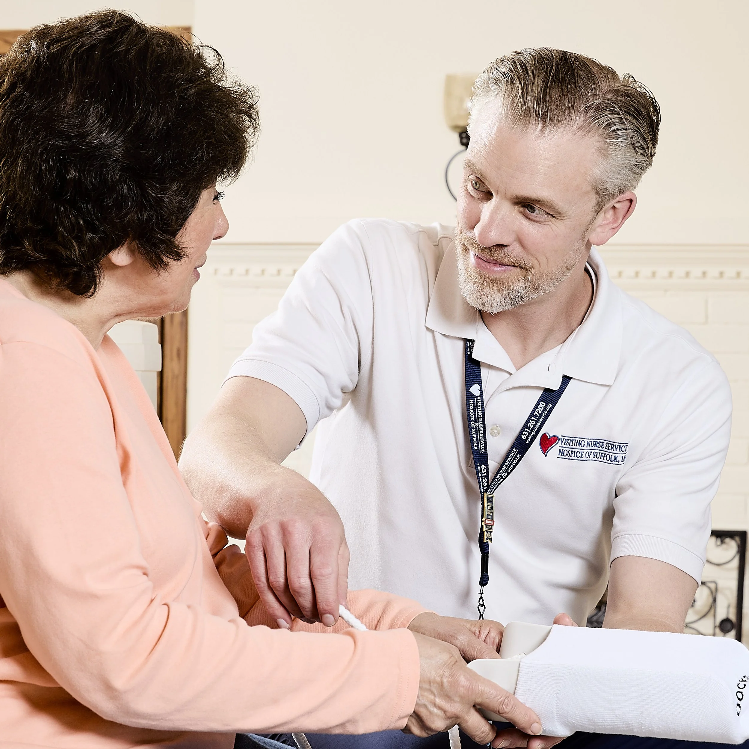 A healthcare worker with a name tag that reads 'Hospice Nurse Service of Suffolk' is assisting an elderly woman in a light orange shirt, holding her arm, in a clinical setting.