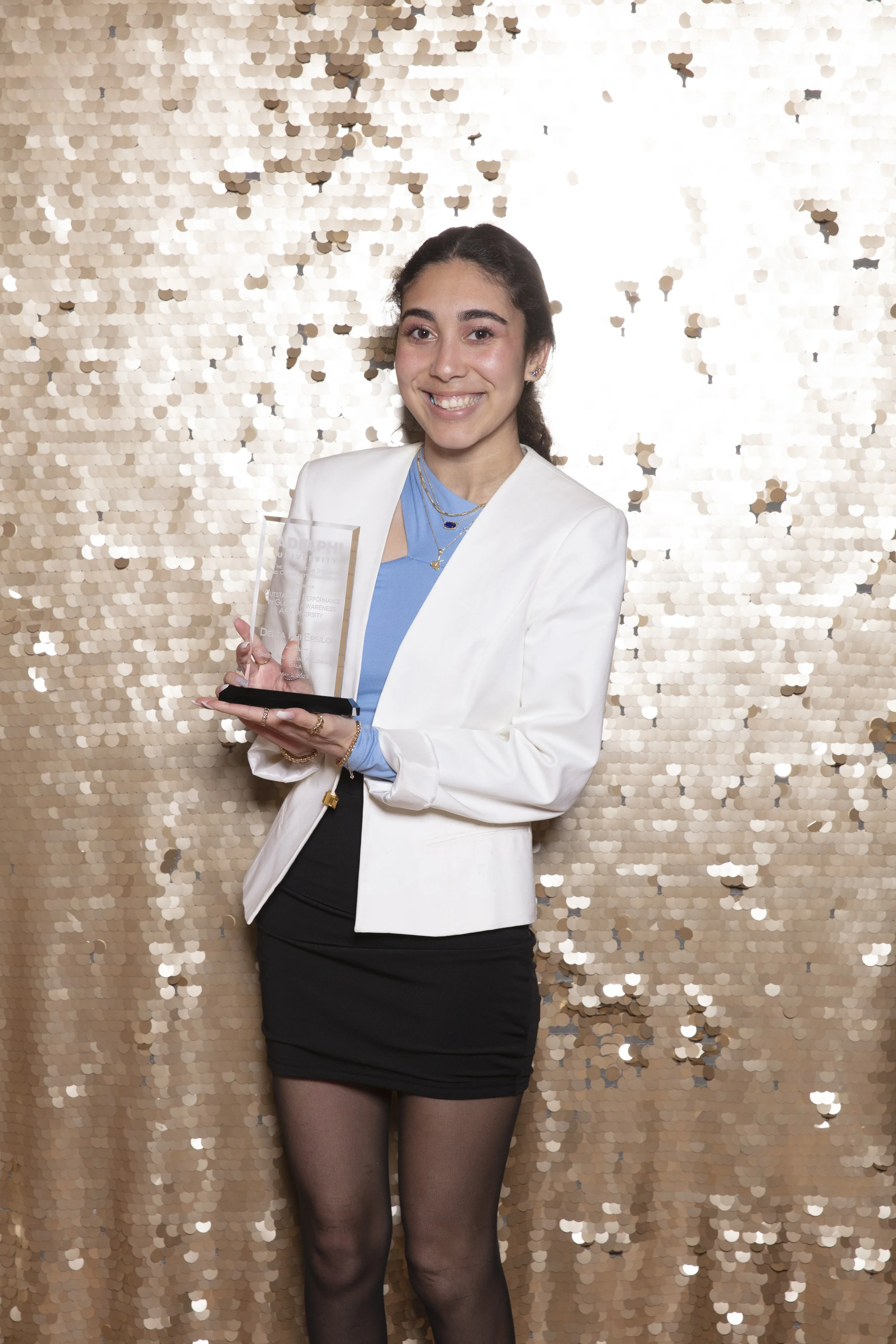 A young woman with dark hair tied back, smiling, dressed in a white blazer, light blue top, black skirt, and black tights, holding a glass award against a gold sequined backdrop.