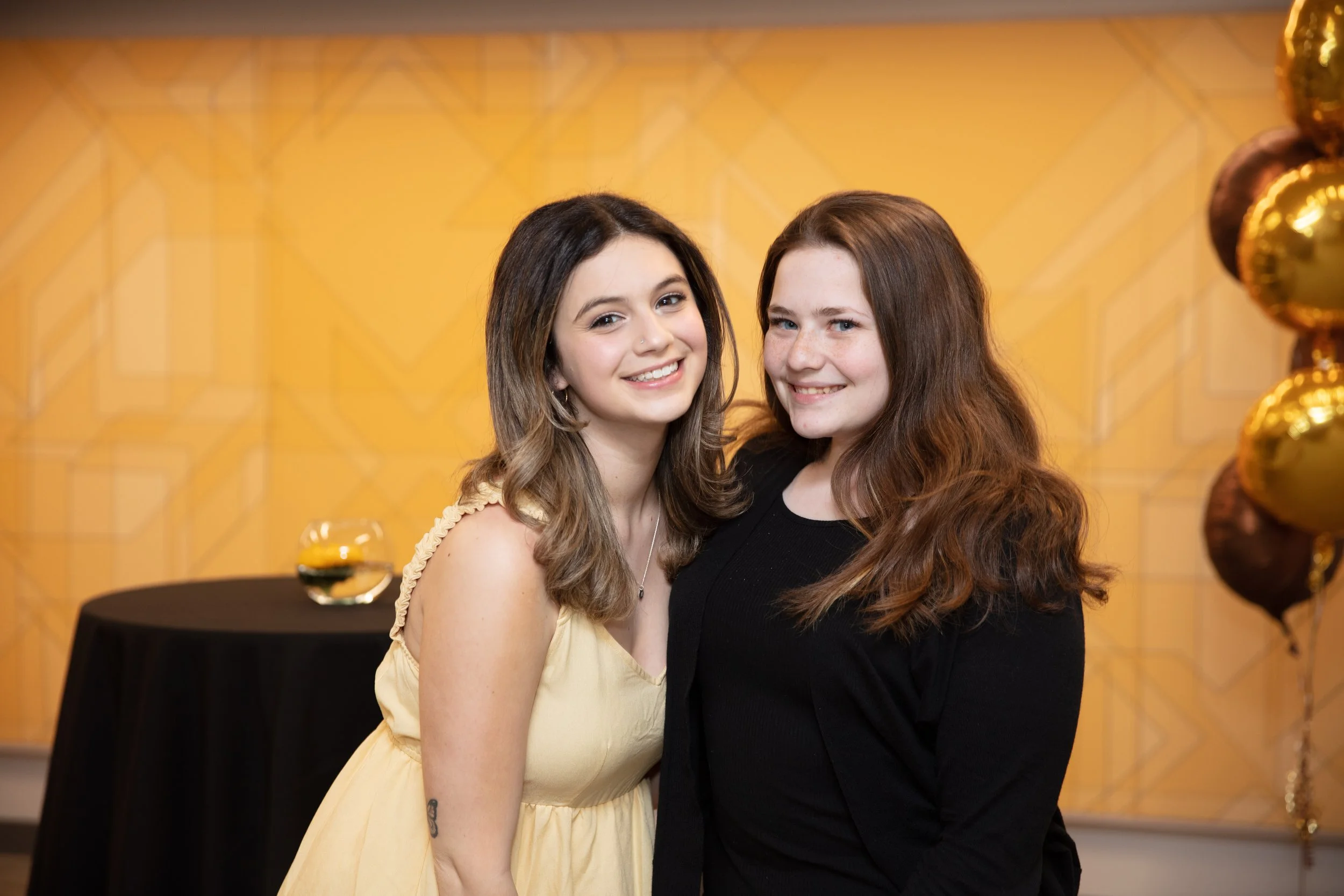 Two young women standing close together, smiling at the camera, at a decorated event with balloons and a gold themed background.