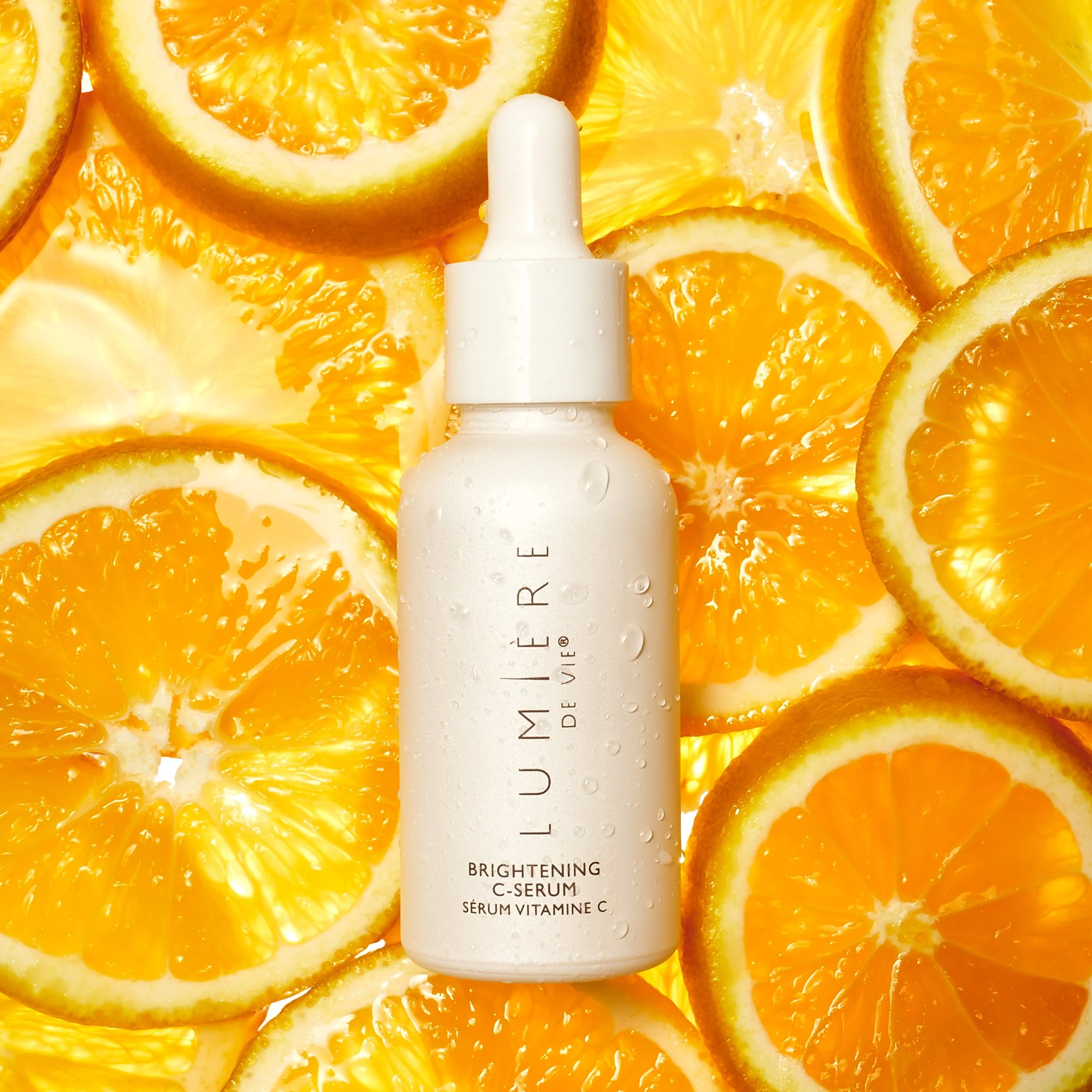 A white bottle of Lumière de Vie Brightening C-Serum surrounded by sliced oranges and lemon slices.