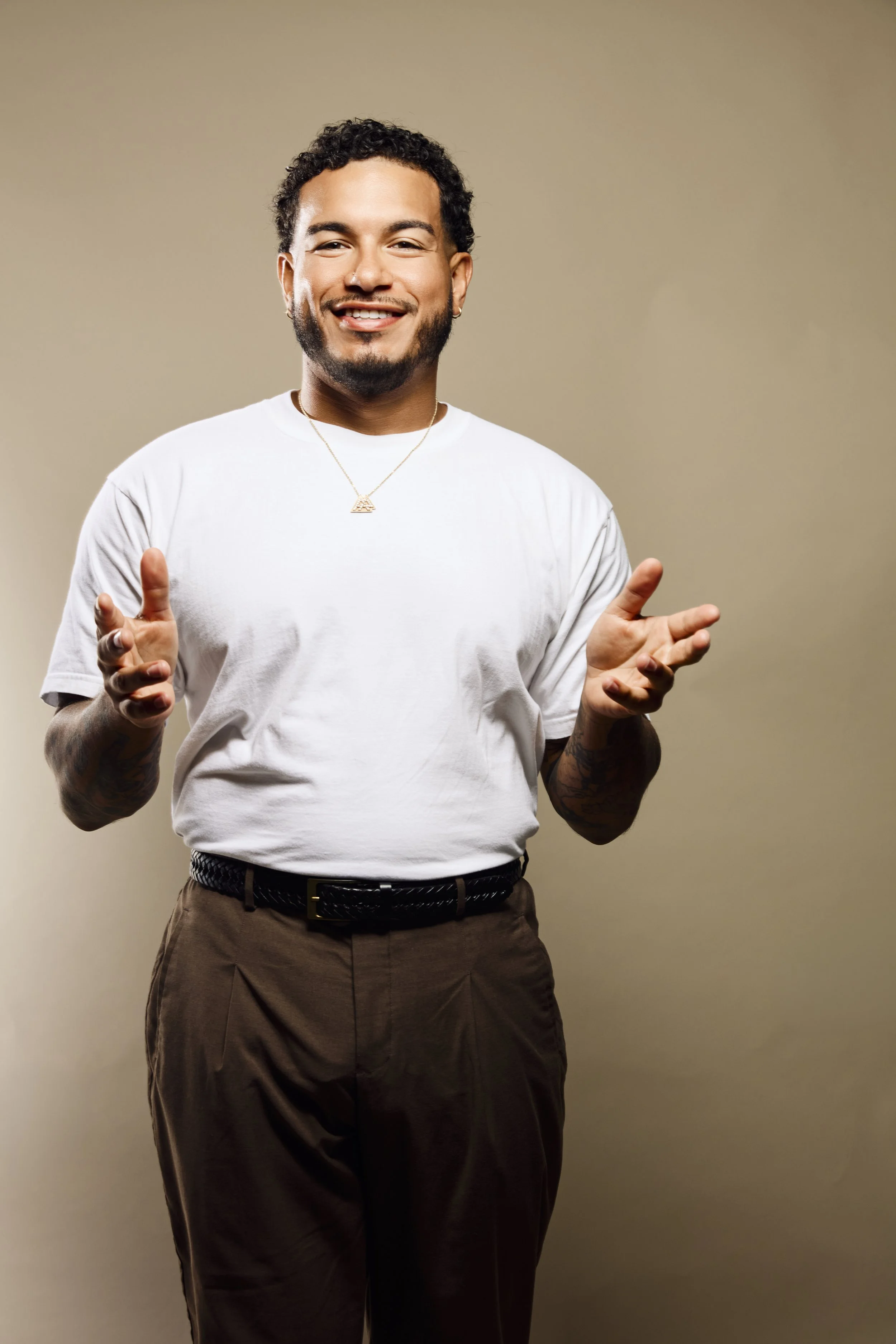 A young man with curly black hair, a beard, and earrings, wearing a white t-shirt, brown belt, and brown pants, stands smiling against a plain beige background. He is gesturing with his hands, palms open and facing upward.