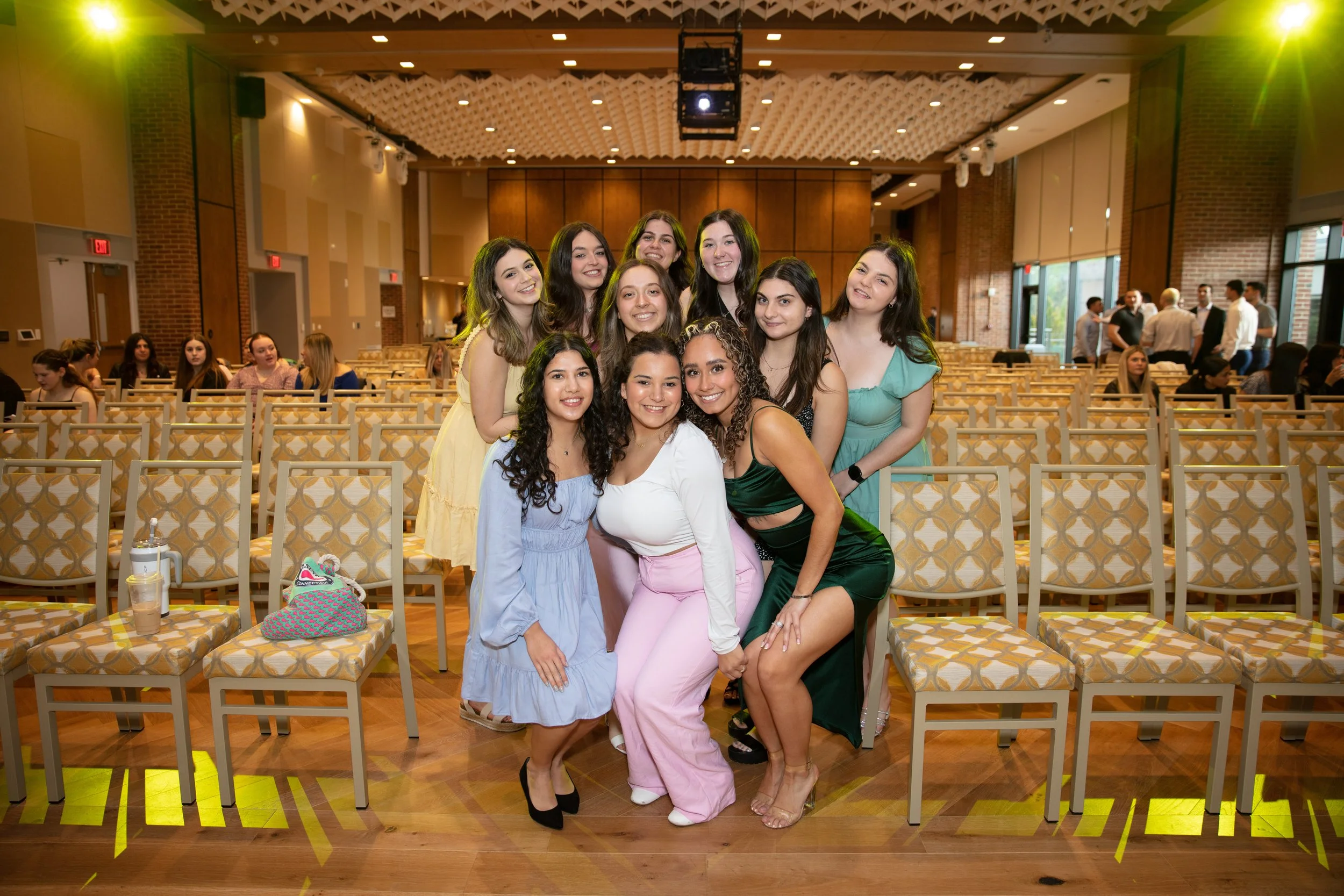 Group of young women posing together at an indoor event, with empty chairs and other people in the background.