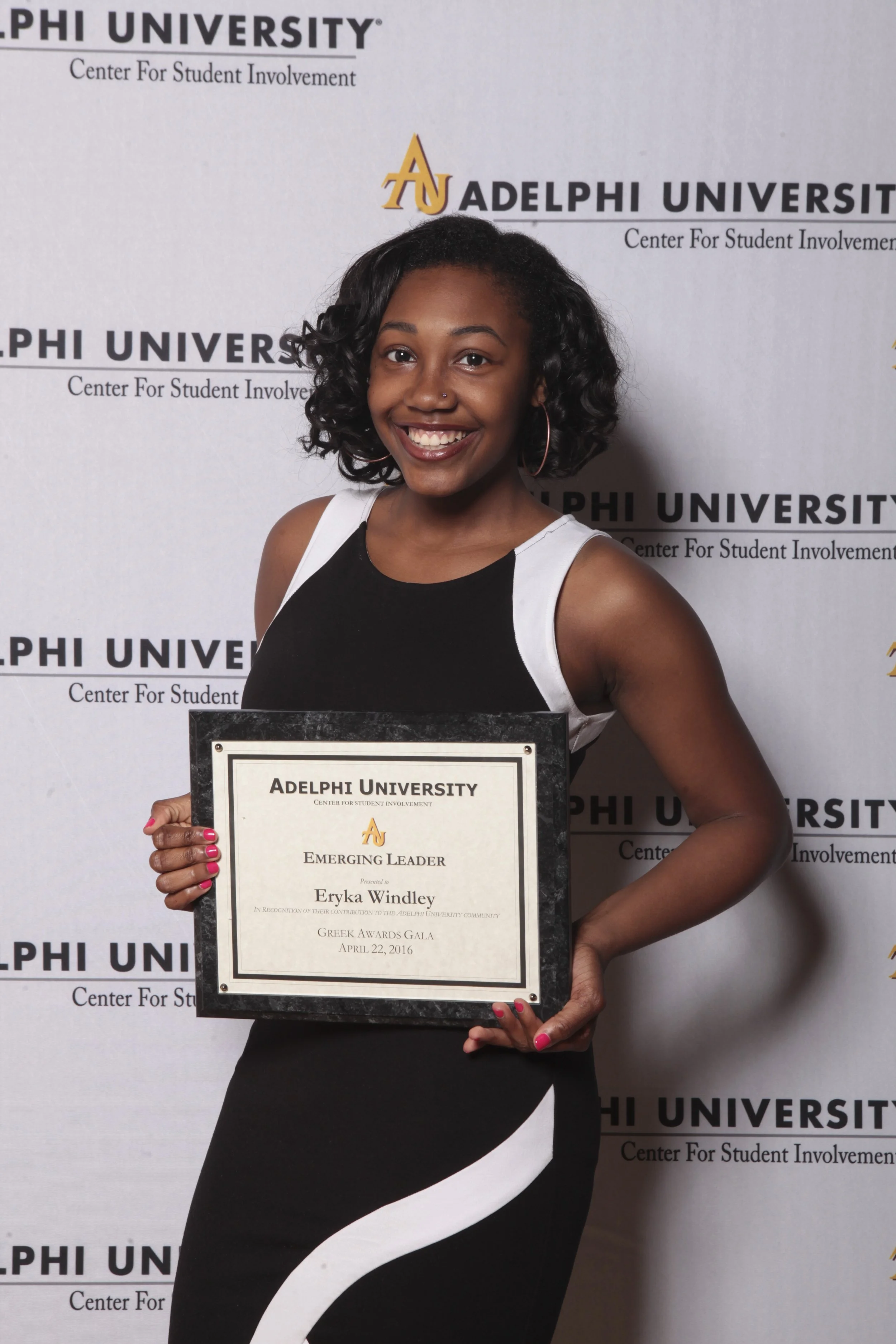 A young woman with curly hair, wearing a black and white dress, smiling and holding a framed certificate in front of a backdrop with 'Adelphi University' and 'Center for Student Involvement' logos.