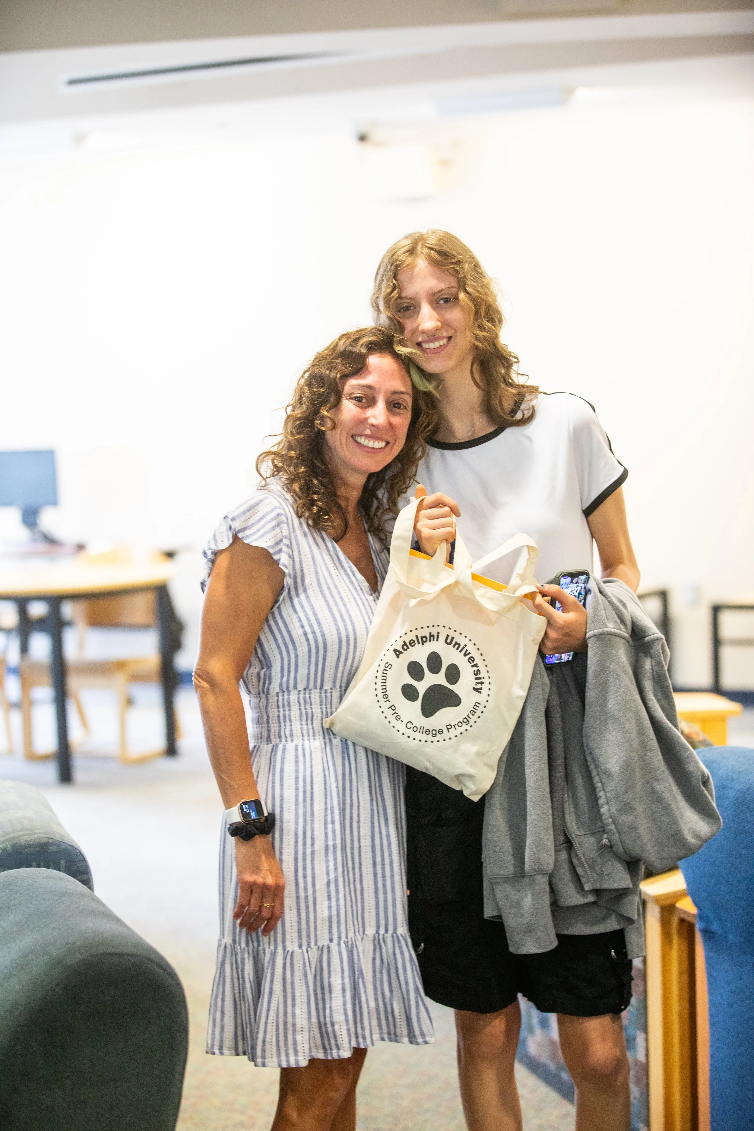 Two women smiling and posing for a photo indoors, holding a tote bag with 'Adelphi University Summer Pre-College Program' and a paw print logo, with a casual background of furniture and computers.