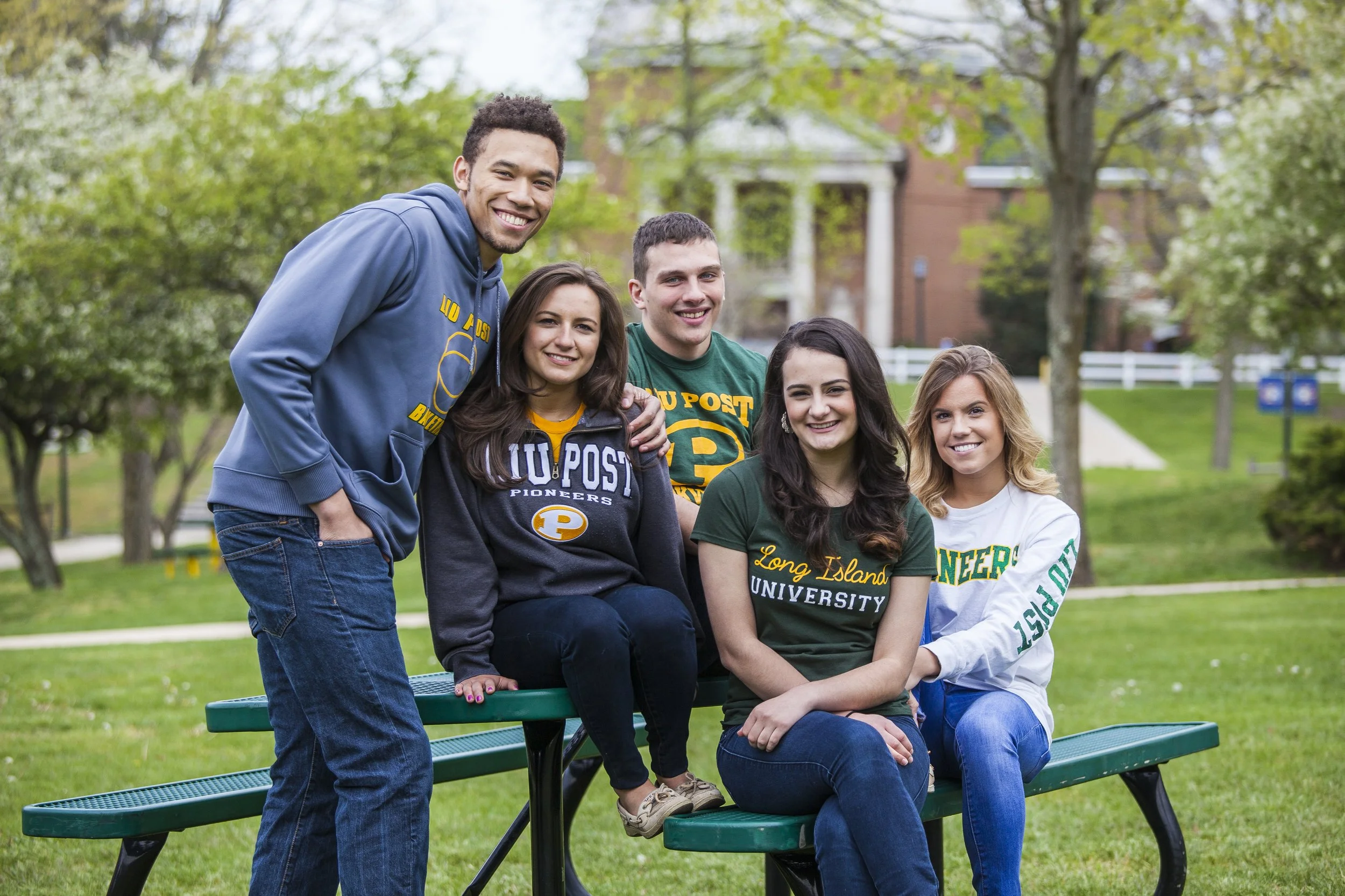Group of five young adults, three women and two men, sitting and standing on a green park bench outdoors, smiling. They are wearing Long Island University apparel. Green grass, trees, and a brick building are in the background.