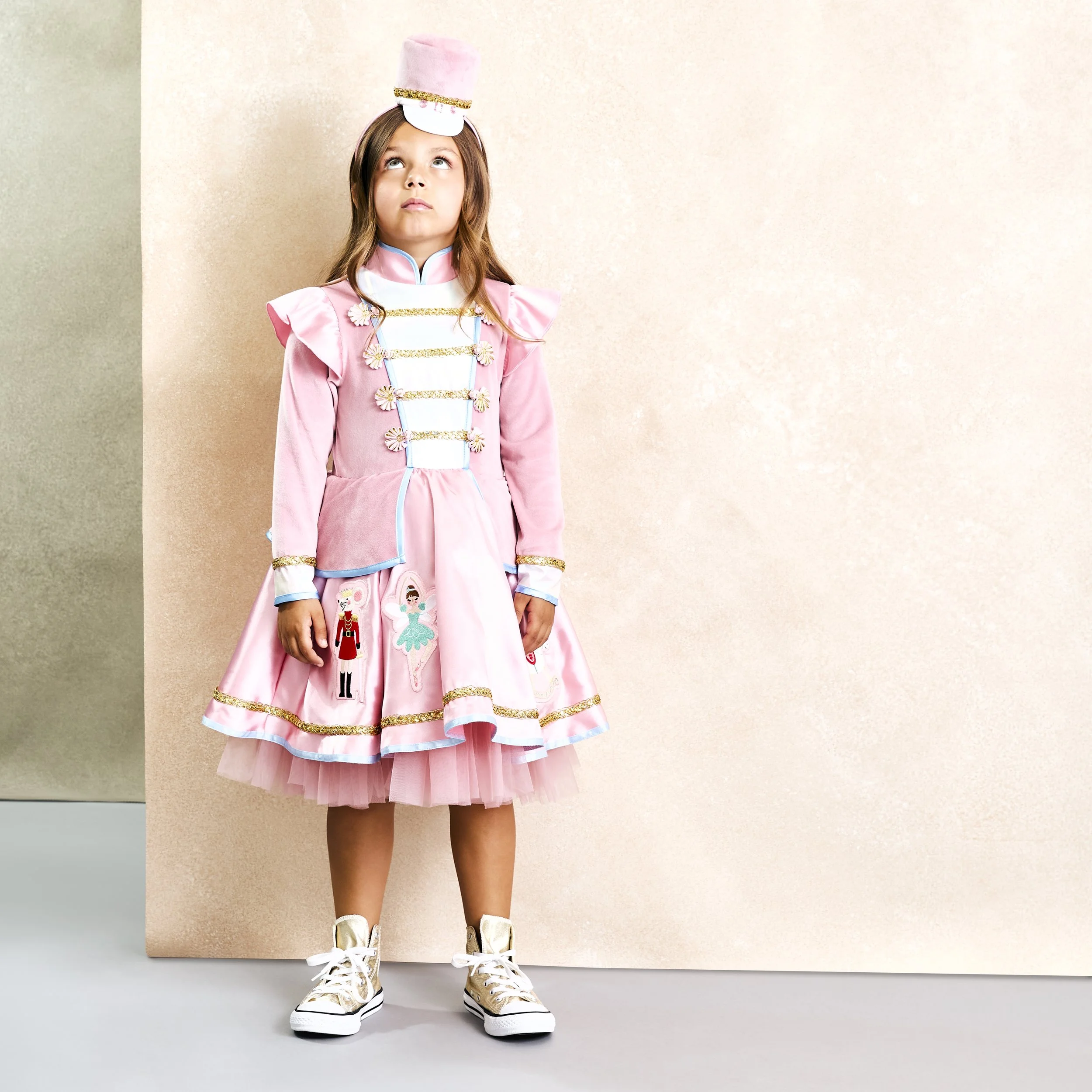 Young girl in a pink, princess-style costume with military details, standing against a peach and beige background, wearing gold high-top sneakers and balancing a pink hat with white and gold accents on her head.