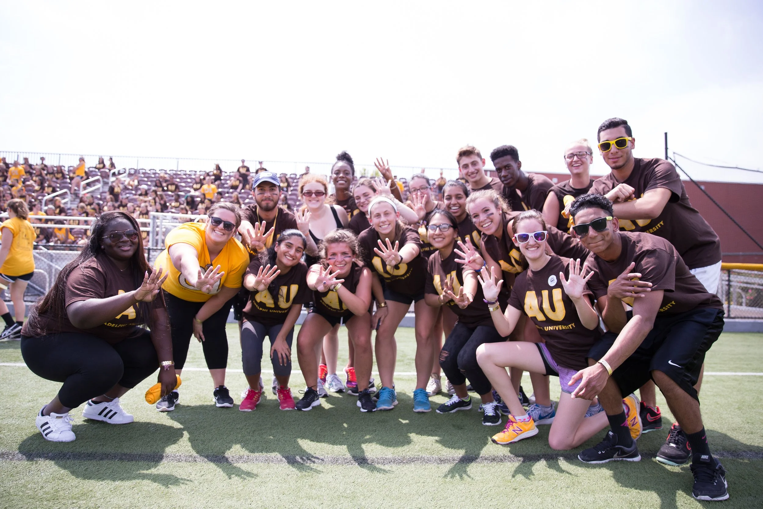 A large group of young people wearing Auburn University shirts, some in sunglasses, smiling, waving, and posing on a sports field during a sunny day.