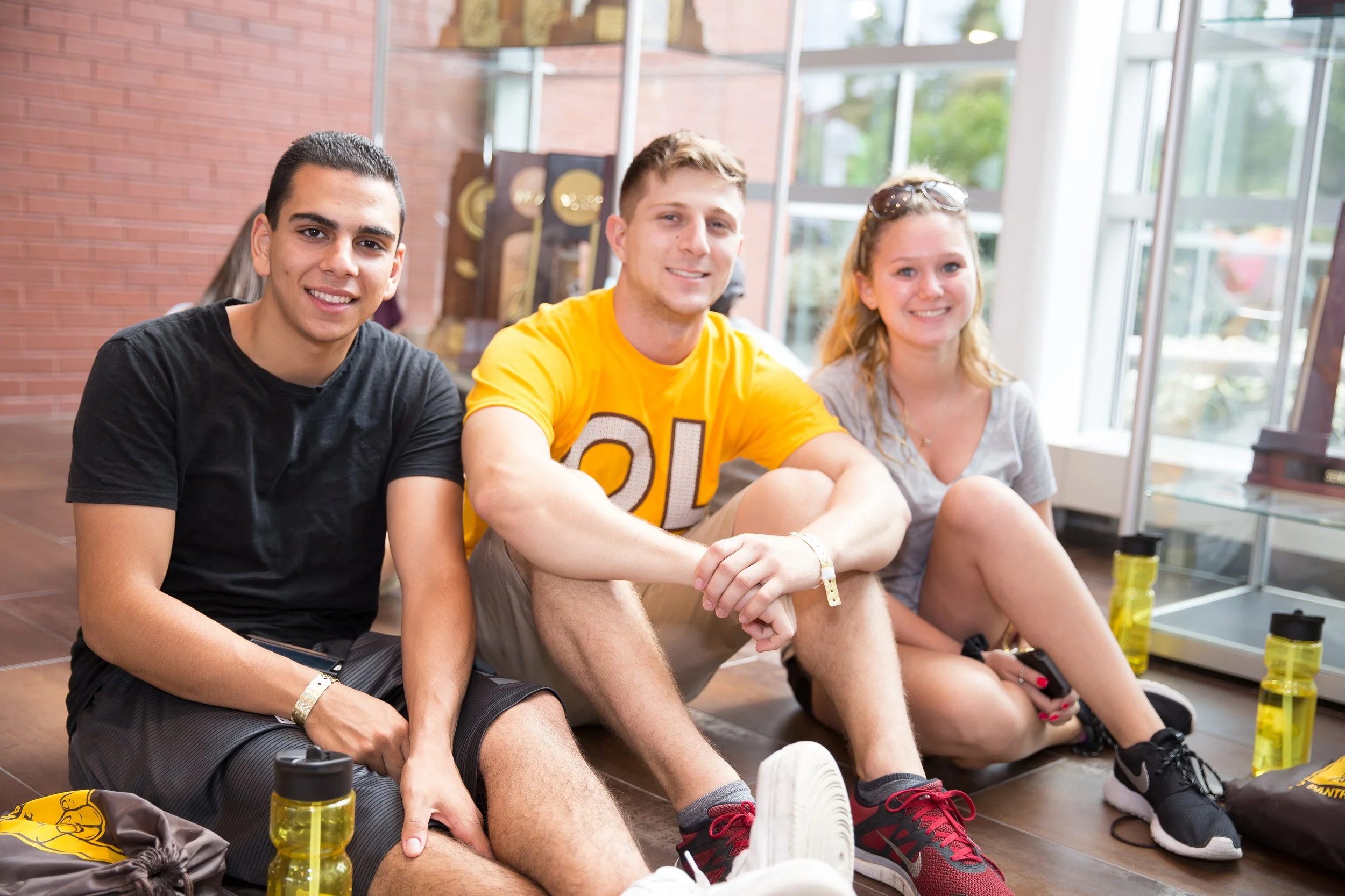 Three young adults sitting on the floor inside a building near a glass display case, smiling at the camera, with water bottles and bags around them.