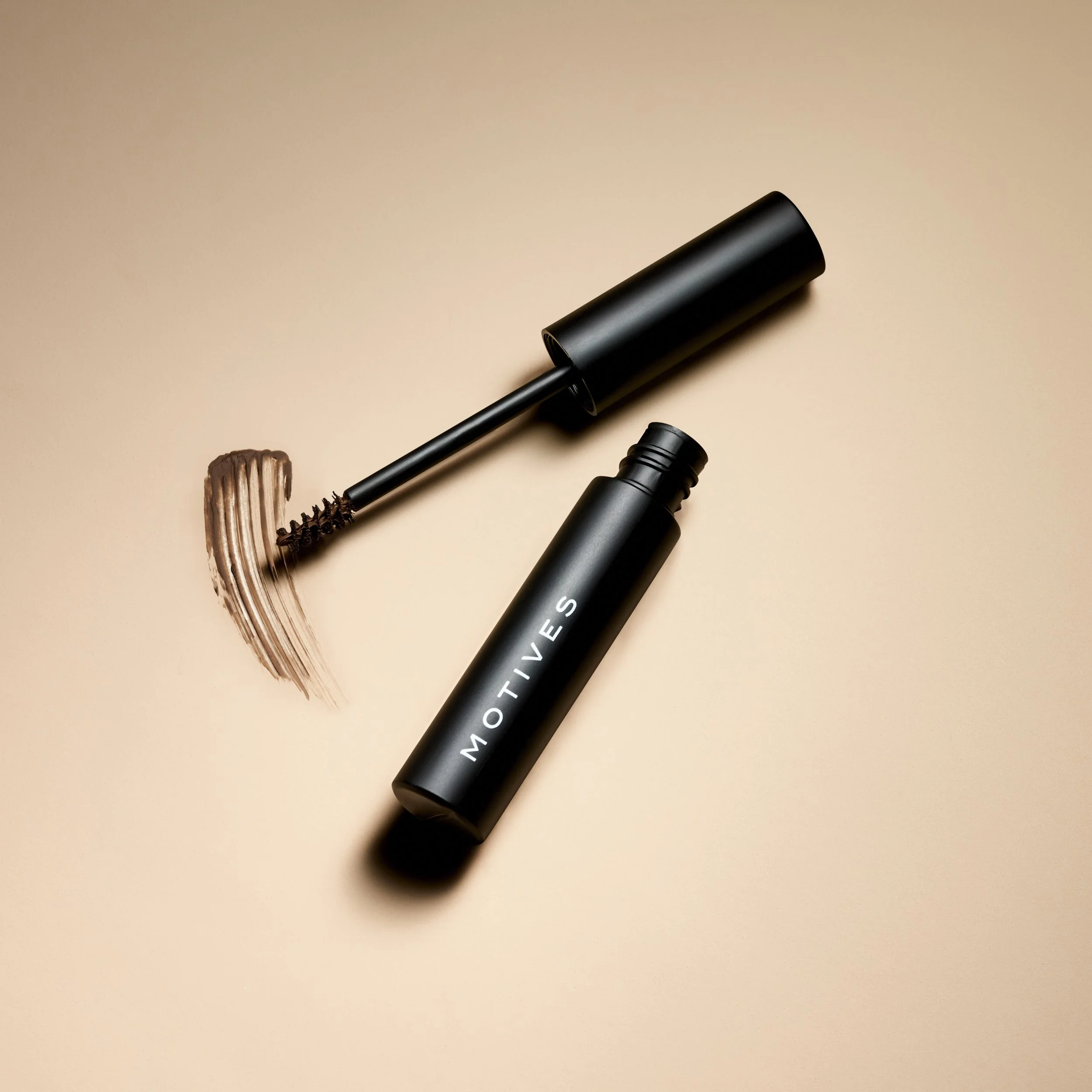 Black mascara tube with the brand name 'MOTIVES' lying on a beige surface, with the mascara wand removed, showing brown mascara on the brush.
