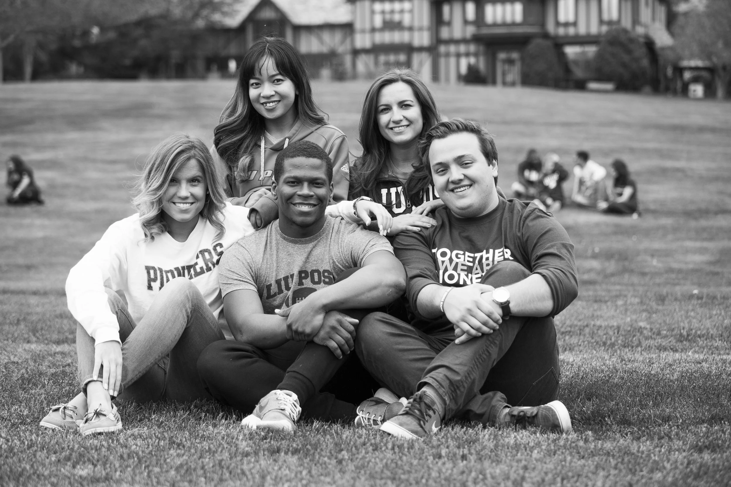 Group of five diverse young people sitting and lying on grass, smiling, with others in the background on a school or park campus.
