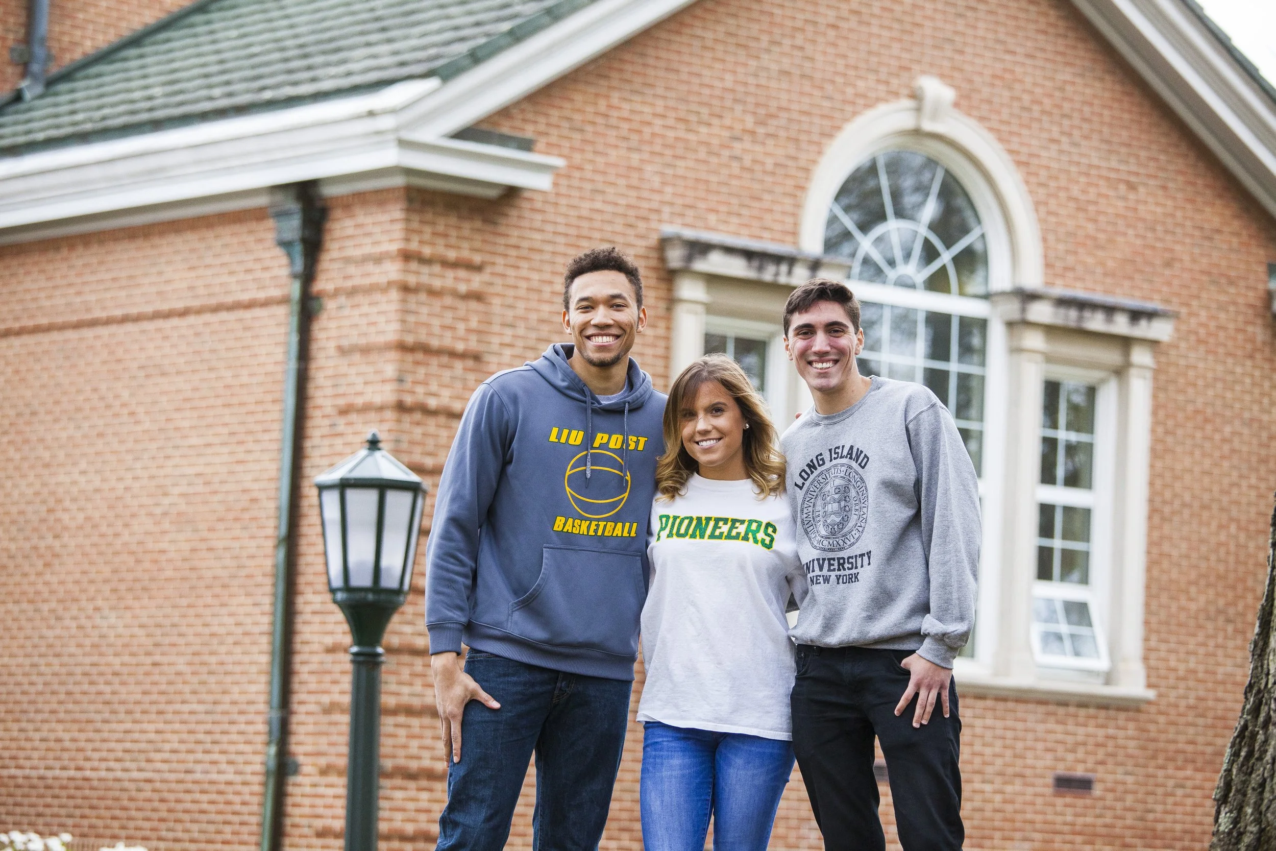 Three young adults standing outside a brick house smiling at the camera. The person on the left wears a gray hoodie with yellow and blue writing. The person in the middle wears a white T-shirt with green lettering. The person on the right wears a gra