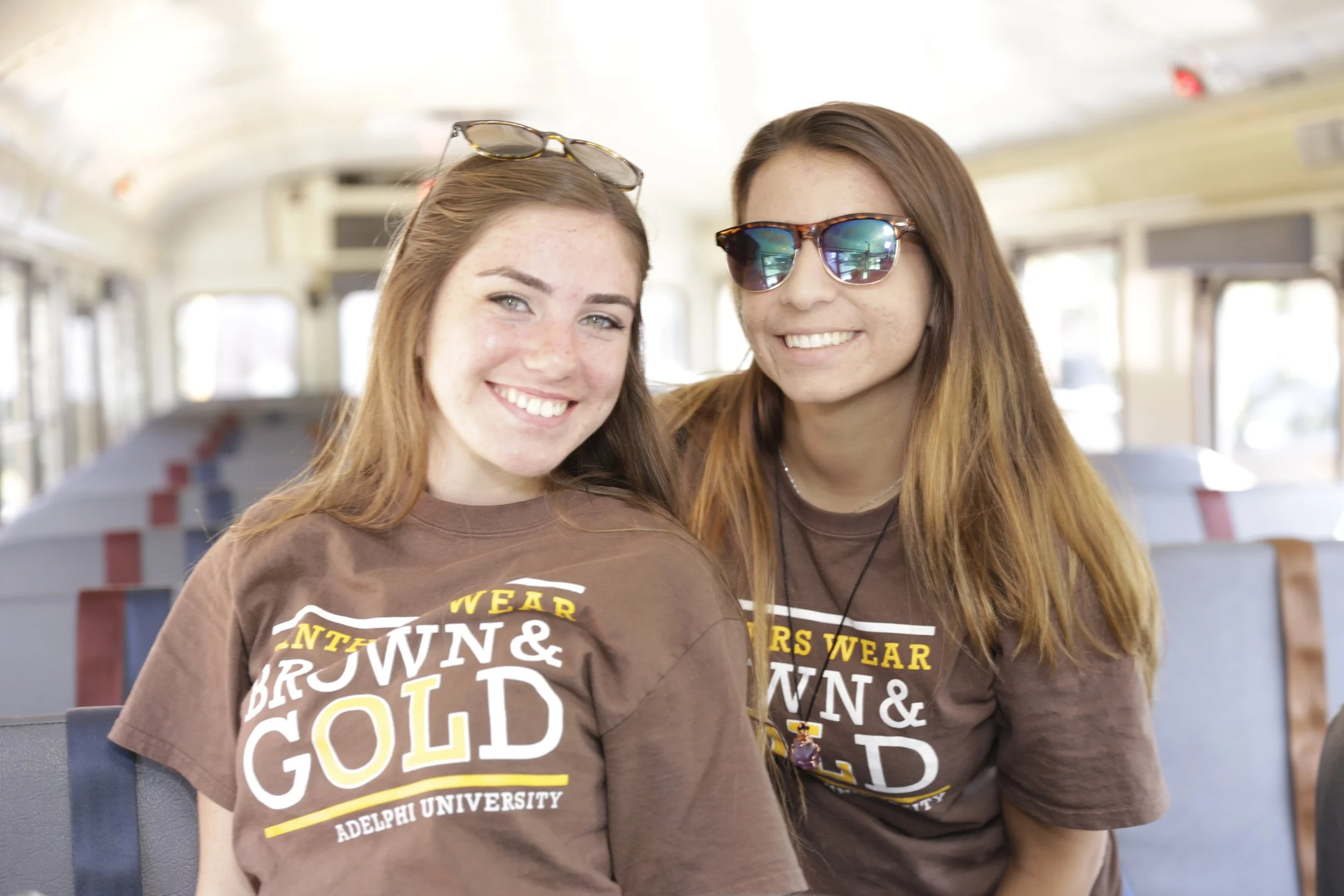 Two young women smiling and sitting together on a school bus. Both are wearing brown t-shirts with yellow and white text that reads 'Brown & Gold, Adelphi University'. They are wearing sunglasses and appear happy.