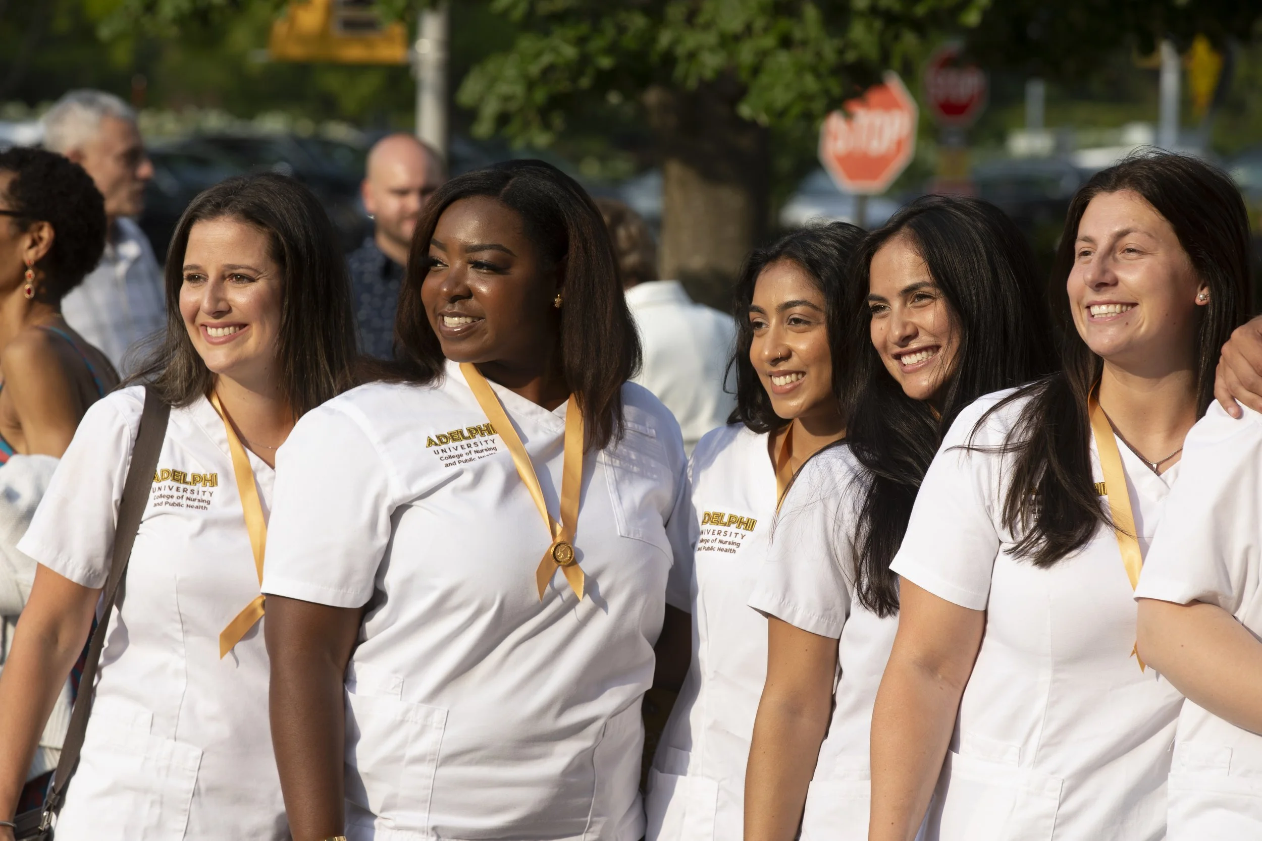 Group of diverse women in white nursing uniforms with medals posing outdoors at a graduation event.