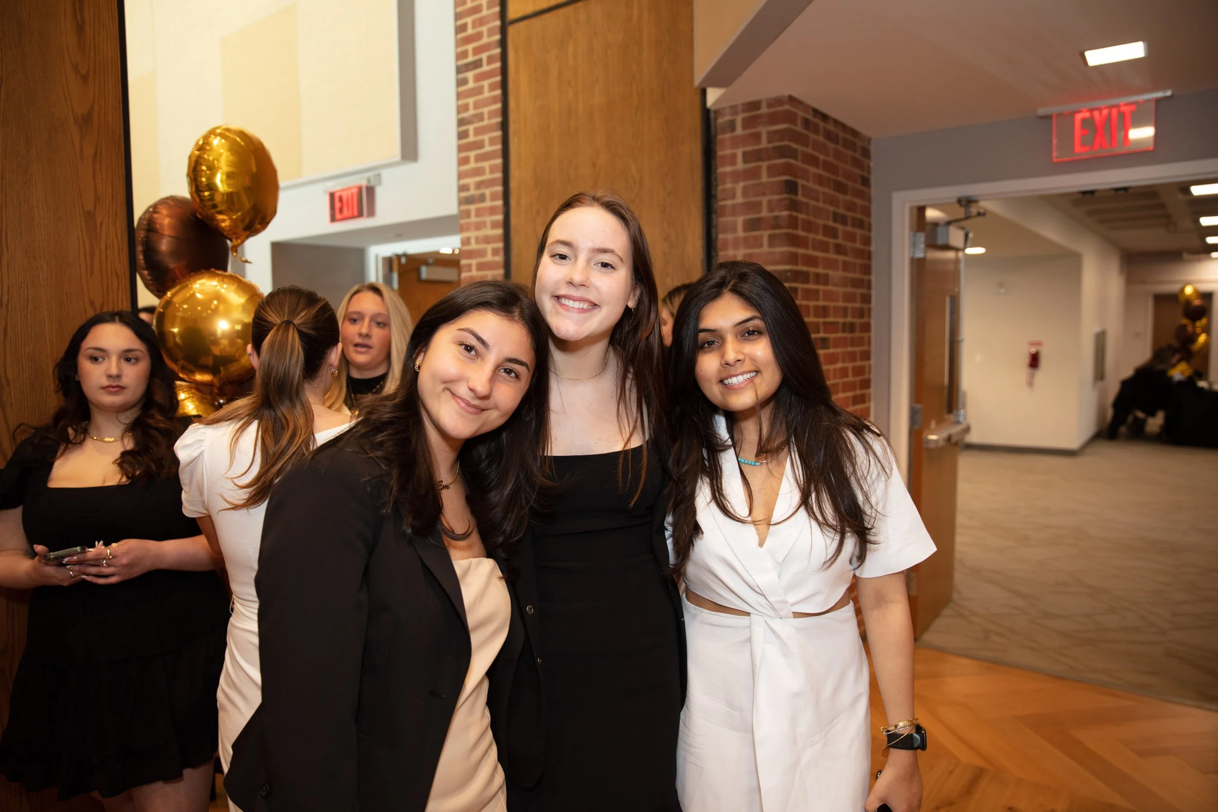 Three young women smiling at the camera at a social event, standing in front of a brick wall with balloons and other people in the background.