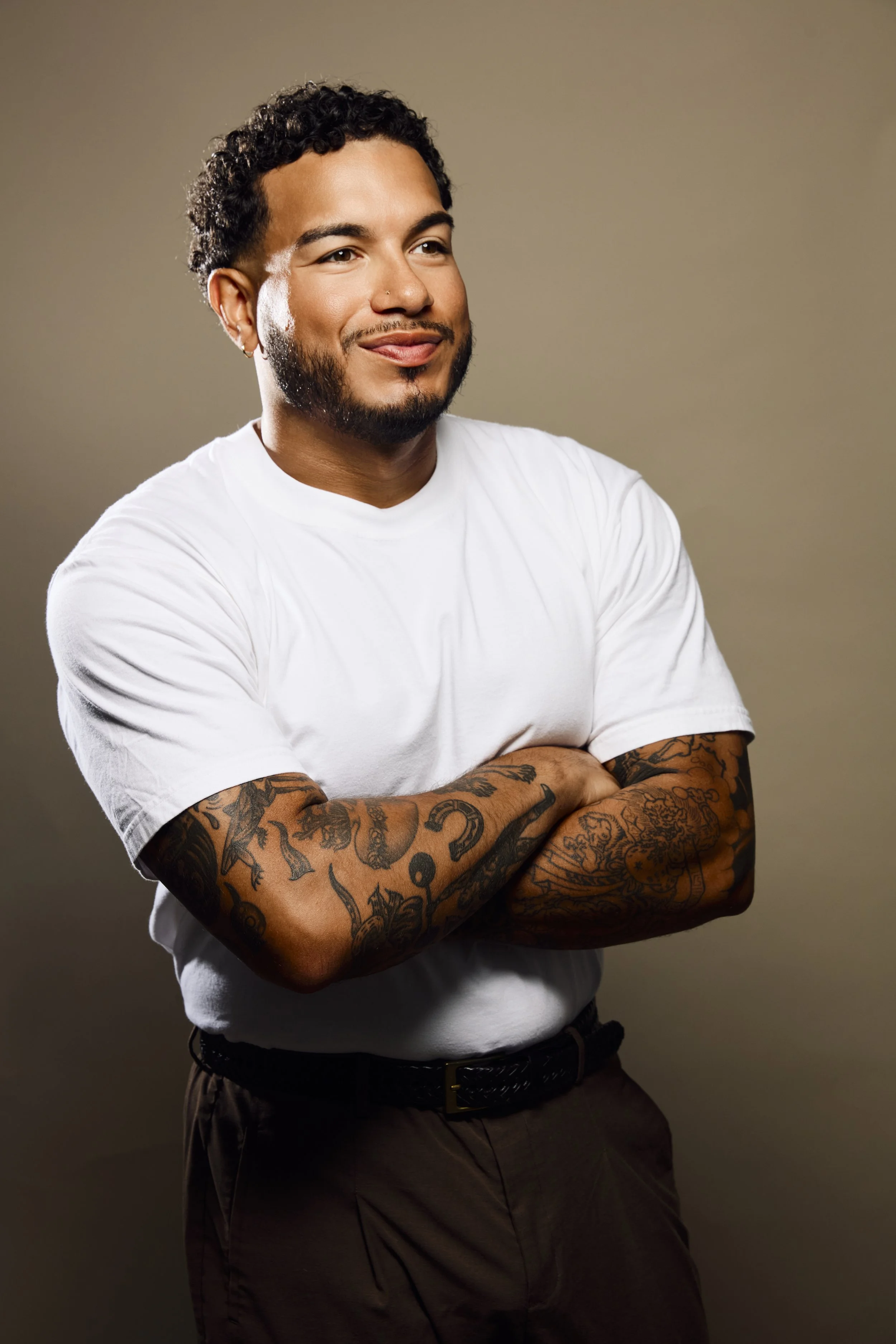 A young man with a beard and tattoos on his arms, wearing a white t-shirt and dark pants with a belt, standing against a plain beige background. He has short curly hair, a small nose ring, and is smiling subtly with his arms crossed.