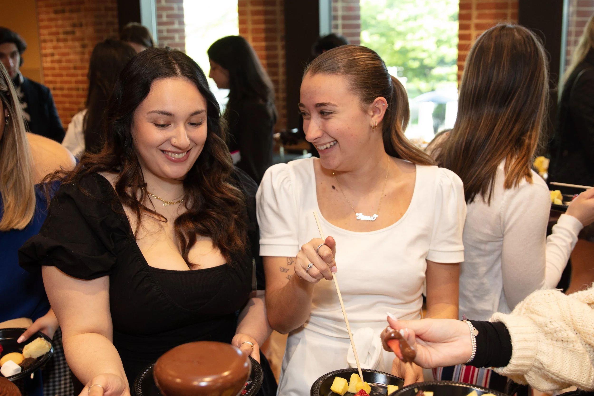 Two young women smiling at each other while holding plates at a social gathering, with other people and a brick wall in the background