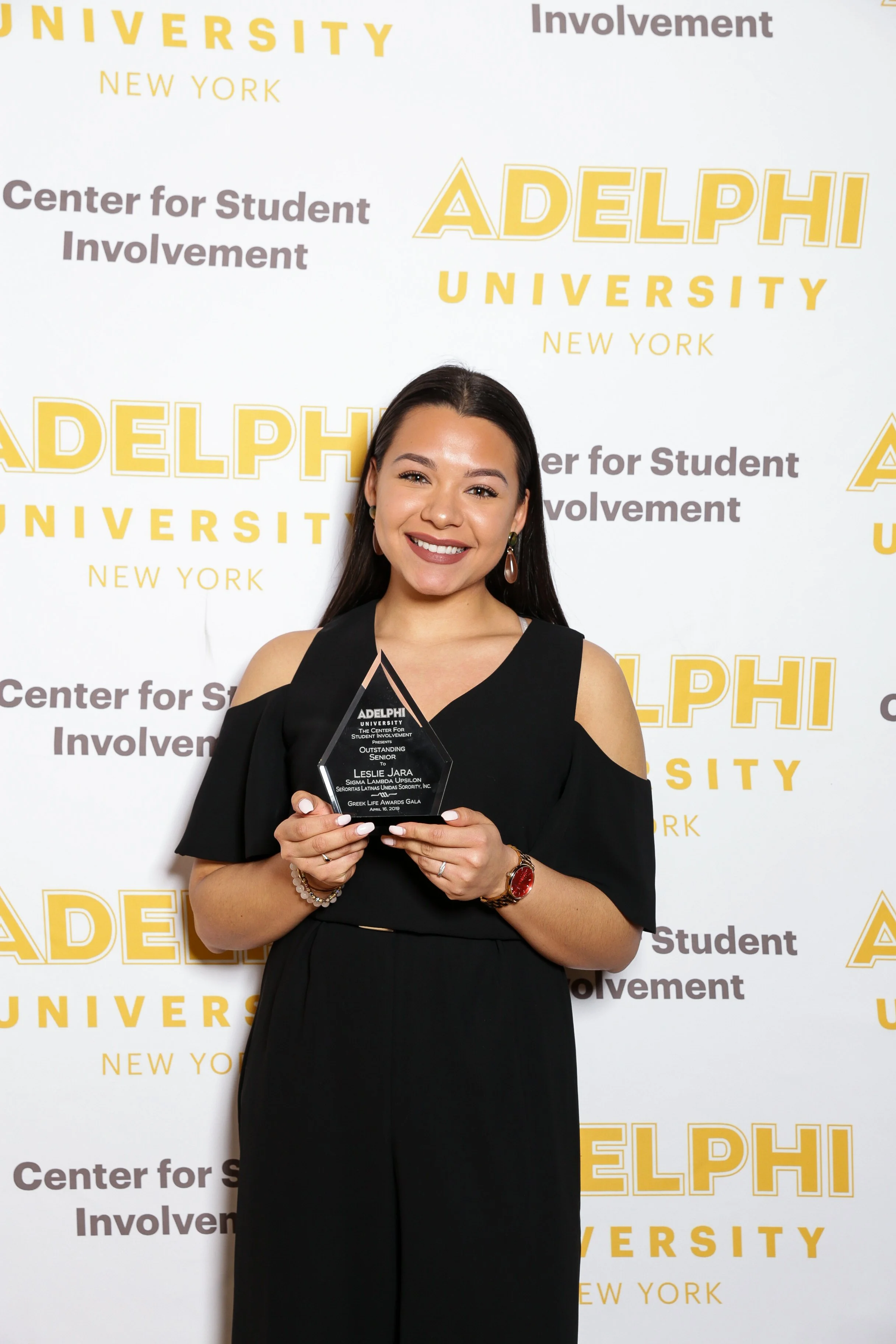 A young woman with long dark hair, wearing a black dress with cold shoulder sleeves, holding a glass award plaque at Adelphi University. She is smiling and standing in front of a backdrop with the Adelphi University logo and the words 'Center for Stu