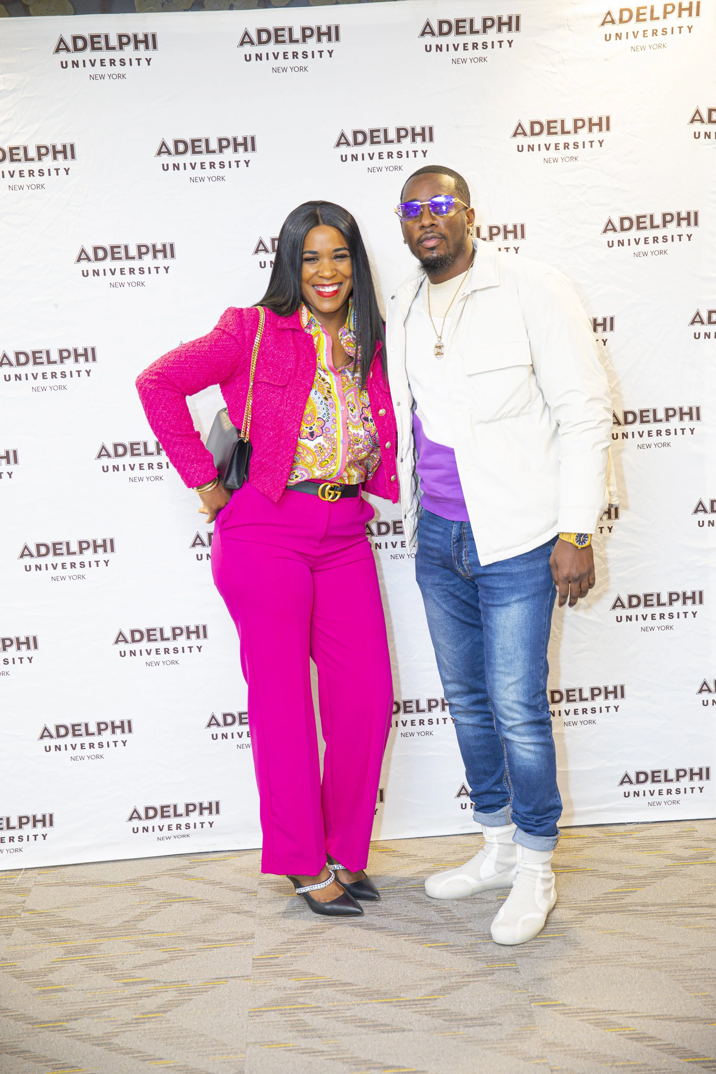 Two well-dressed individuals, a woman in a bright pink pantsuit and a man in jeans, a white jacket, purple shirt, and white sneakers, standing side by side in front of a step-and-repeat backdrop with "Adelphi University New York" written repeatedly.