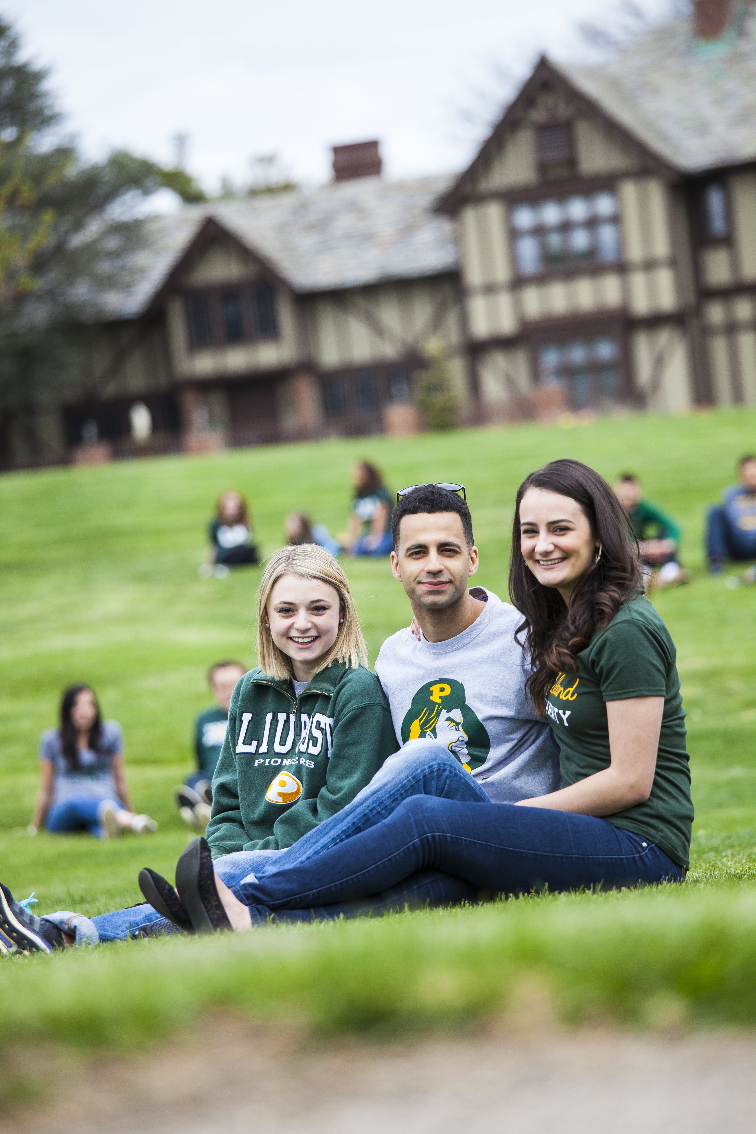 Three young adults sitting on grass in front of a historic house, smiling at the camera, with more people and a house in the background.