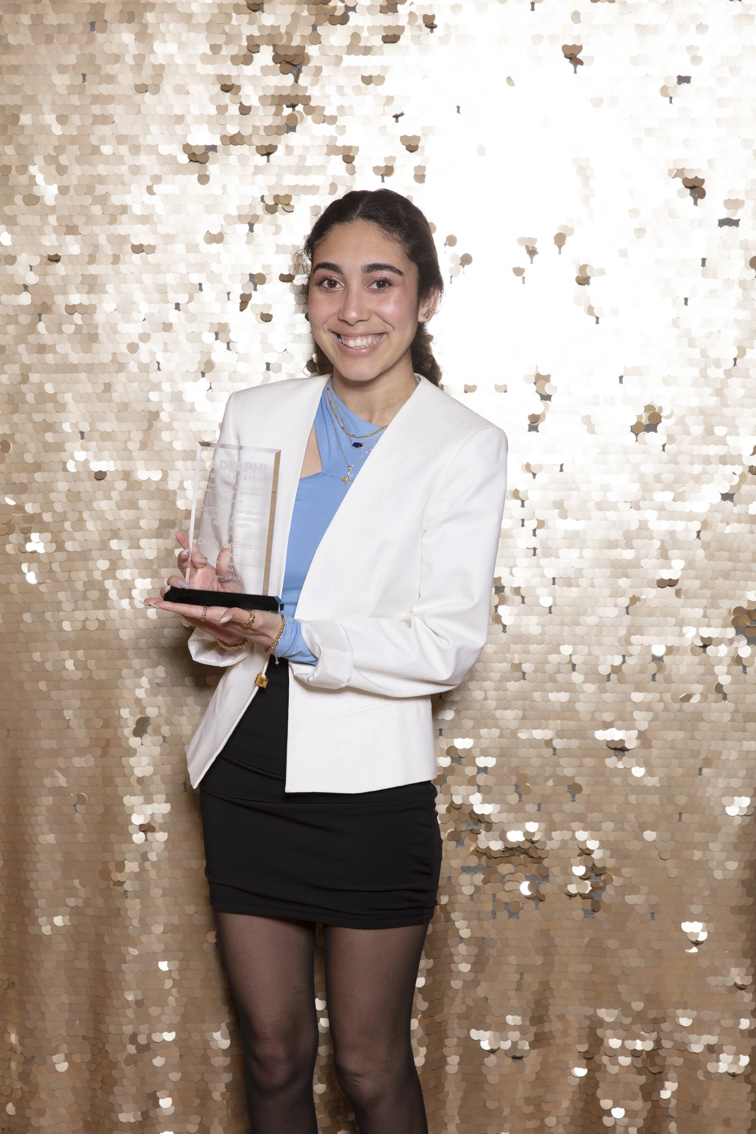 A young woman in a white blazer, blue top, black skirt, and sheer black tights is holding a clear award plaque, smiling against a gold sequin backdrop.