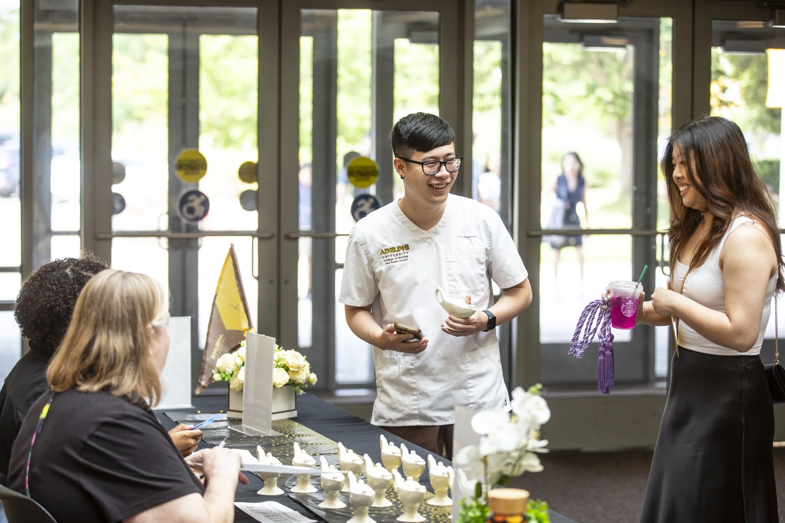 People interacting at a welcoming event, with a woman holding a pink drink and a man in white attire smiling at her, in a well-lit indoor space near glass doors.