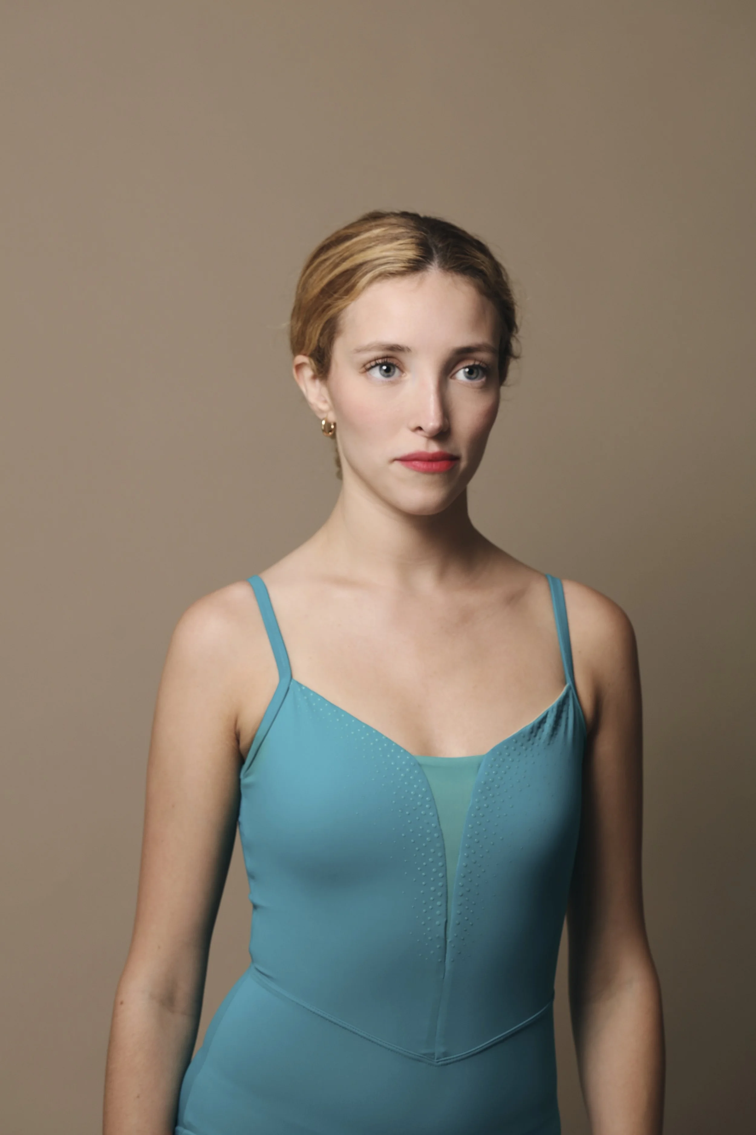 A woman with short, reddish-brown hair and light skin wearing a blue sleeveless top with thin straps against a plain brown background.
