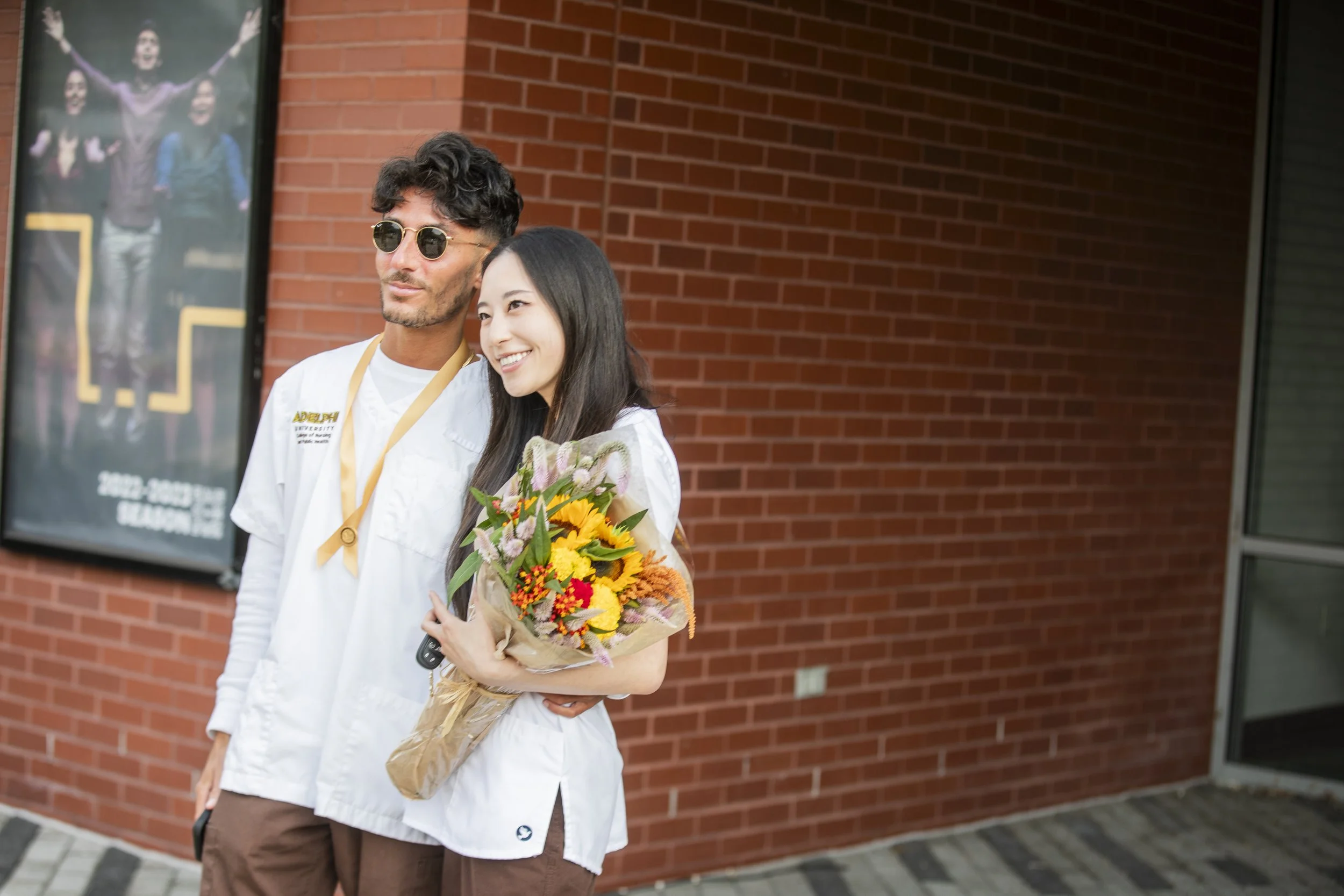 A young woman with long dark hair smiling and holding a colorful bouquet of flowers, standing beside a man with sunglasses and a white jacket, both smiling outside near a brick building.