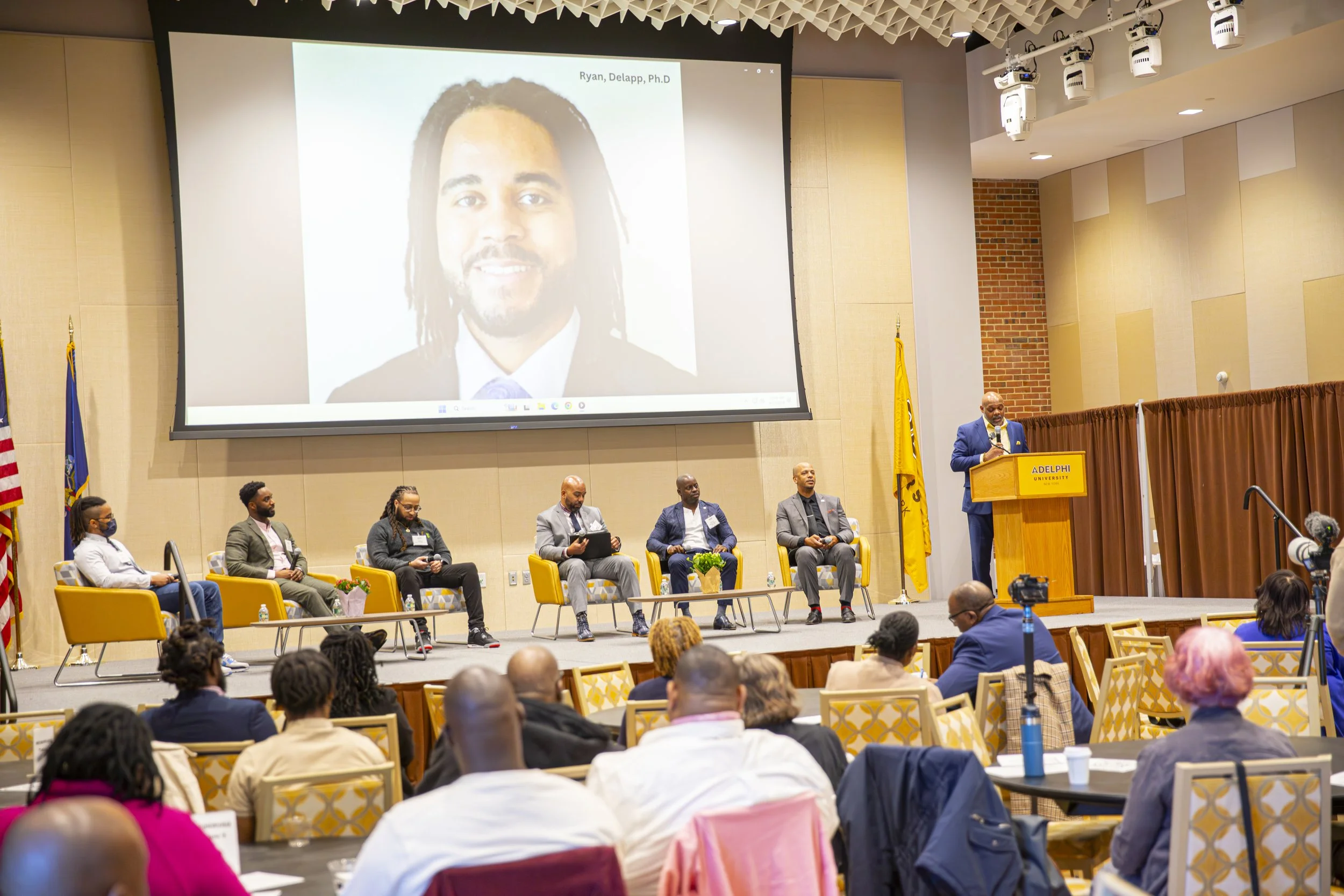 Indoor conference with a panel of six people seated on stage, one man at a podium giving a speech, and a large screen displaying an image of a man's face behind the stage. The room has about 20 audience members seated at tables.