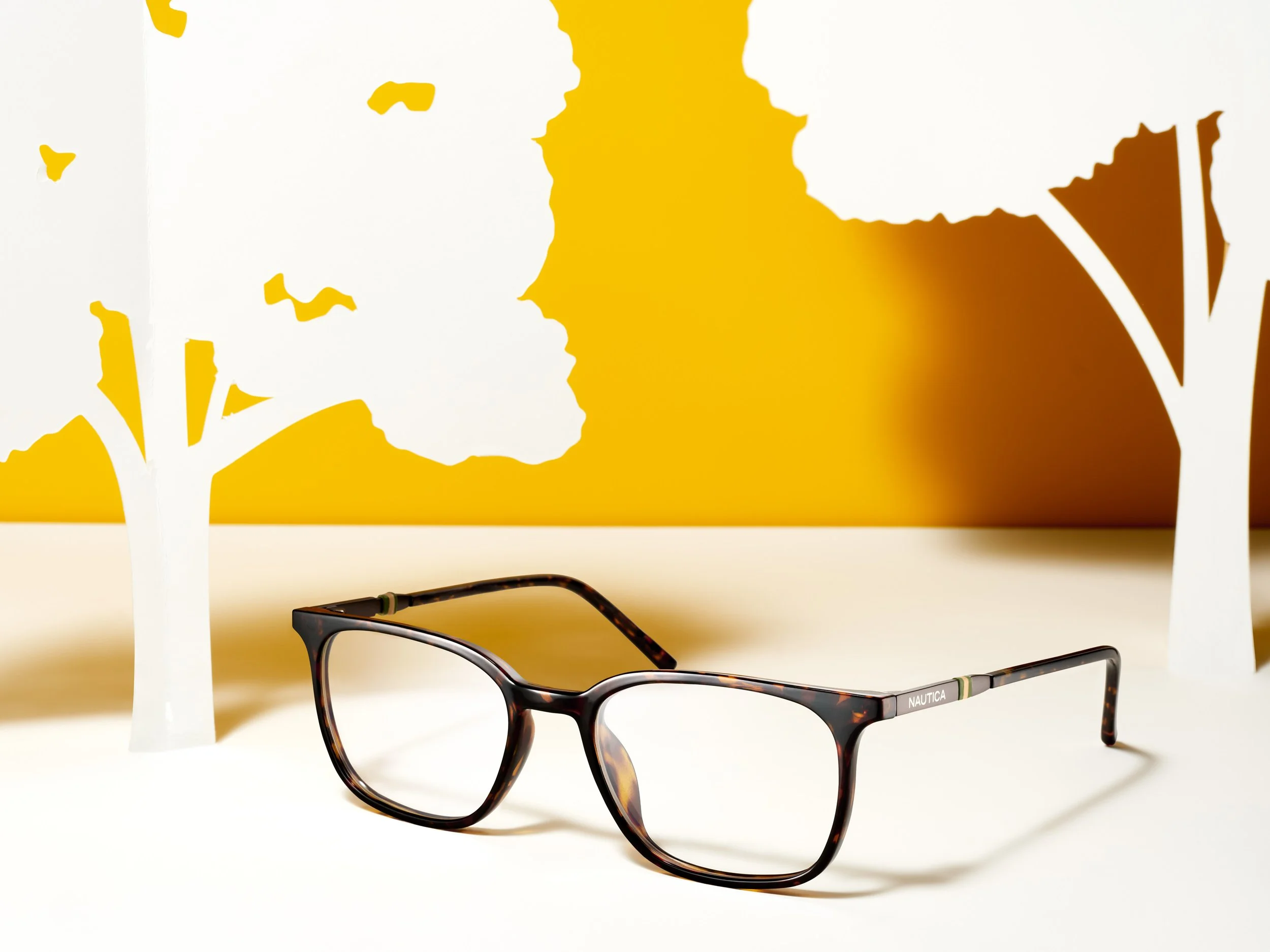 Tortoiseshell eyeglasses with a black and brown pattern placed on a white surface, with cutout paper trees and an orange background in the scene.