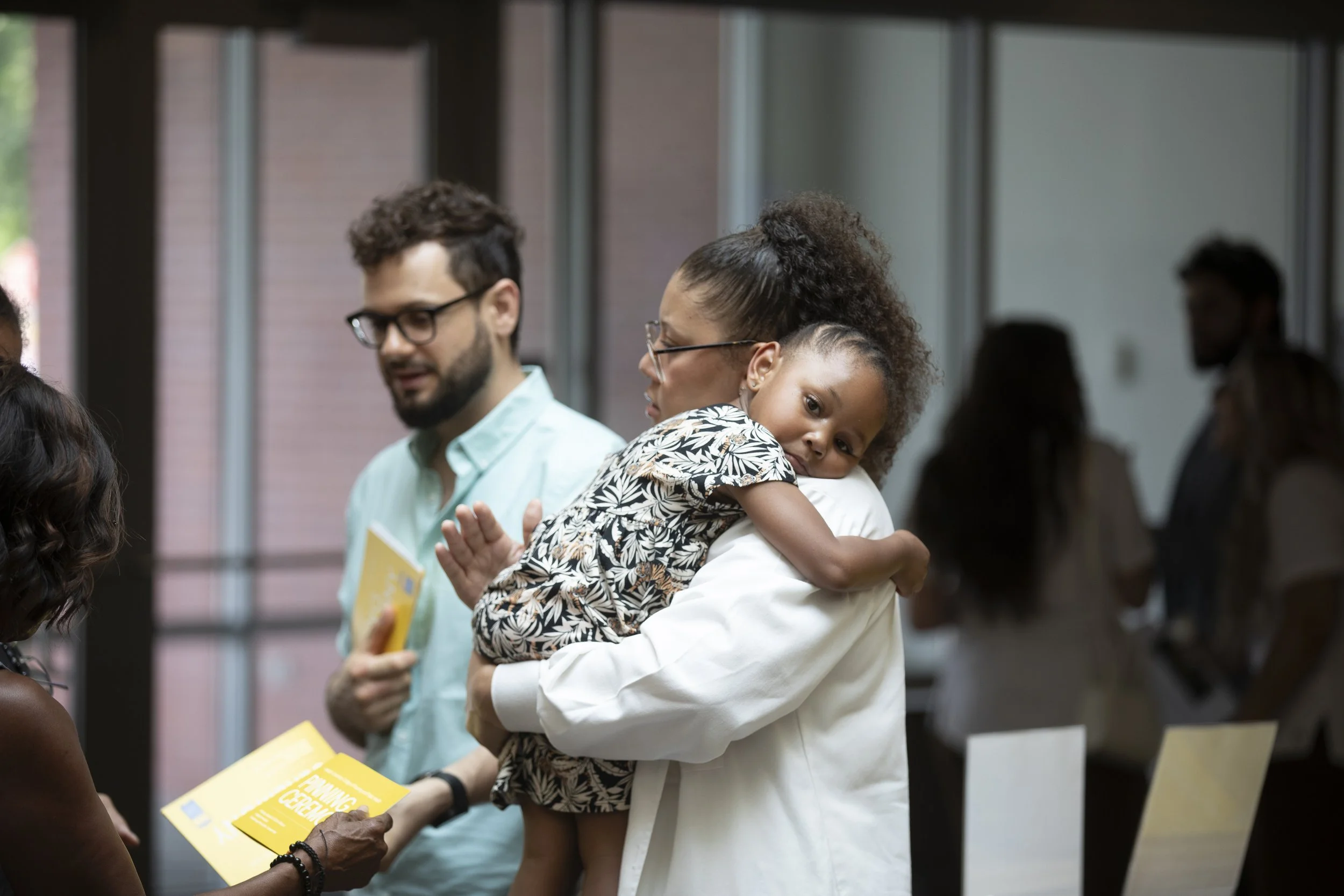 A woman holding a young girl in her arms at an indoor event. The girl is resting her head on the woman's shoulder, and they are surrounded by other people.