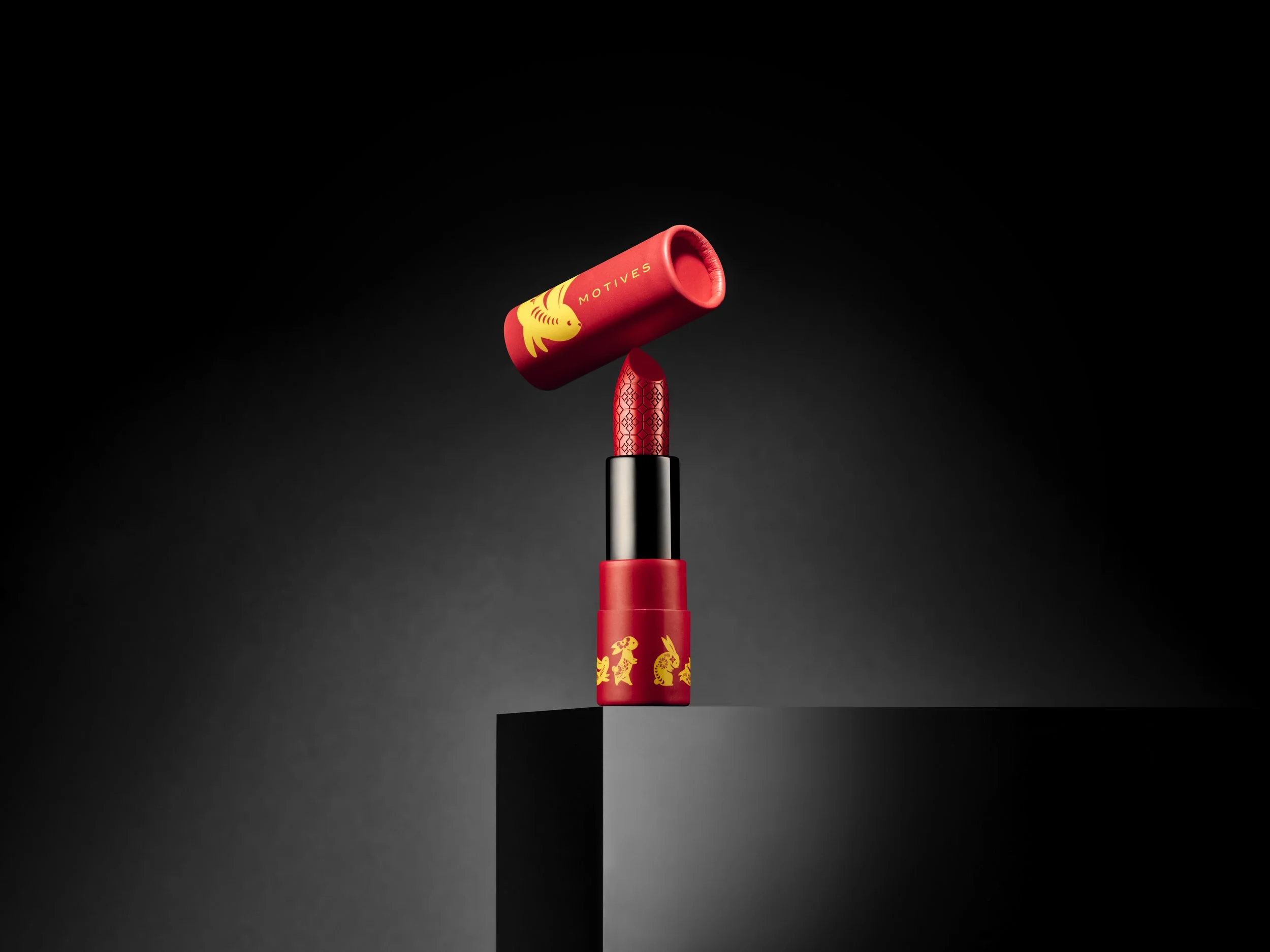 A lipstick with a red casing decorated with yellow animals and mushrooms stands on a black block, with the cap balanced on the lipstick in a black background.
