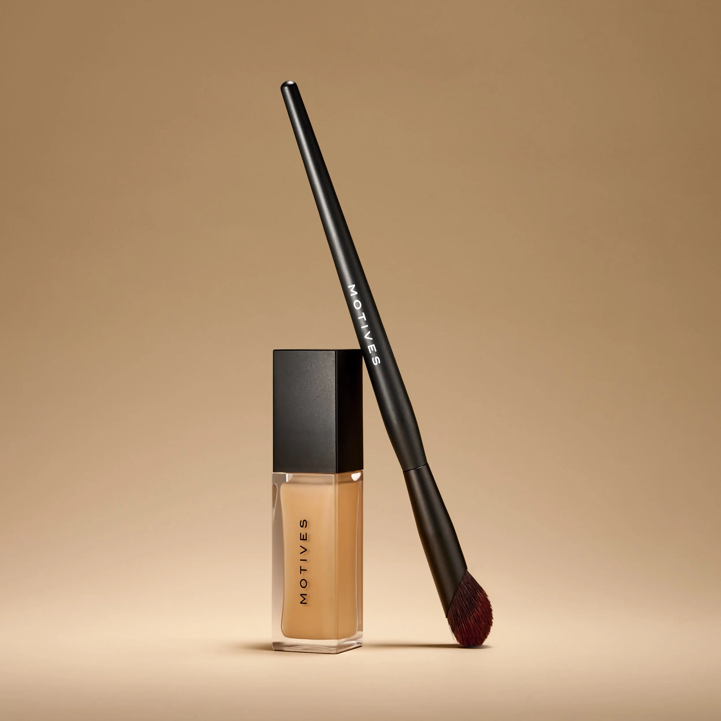 Beauty products including a bottle of beige liquid foundation with a black cap and an angled makeup brush with dark red bristles and a black handle labeled MOTIVES, against a gradient beige background.