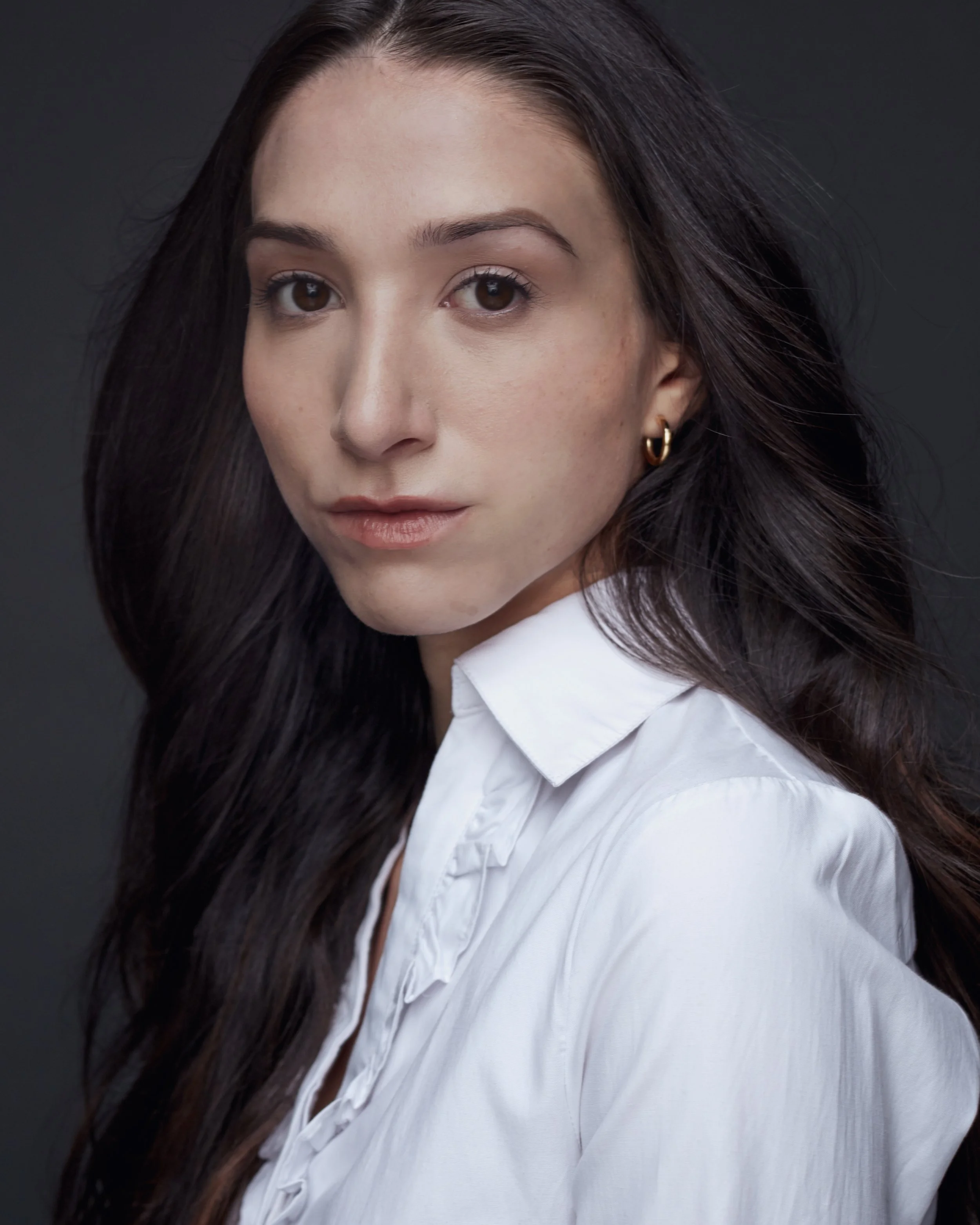 Portrait of a woman with dark brown hair wearing a white blouse and gold earrings against a dark background.